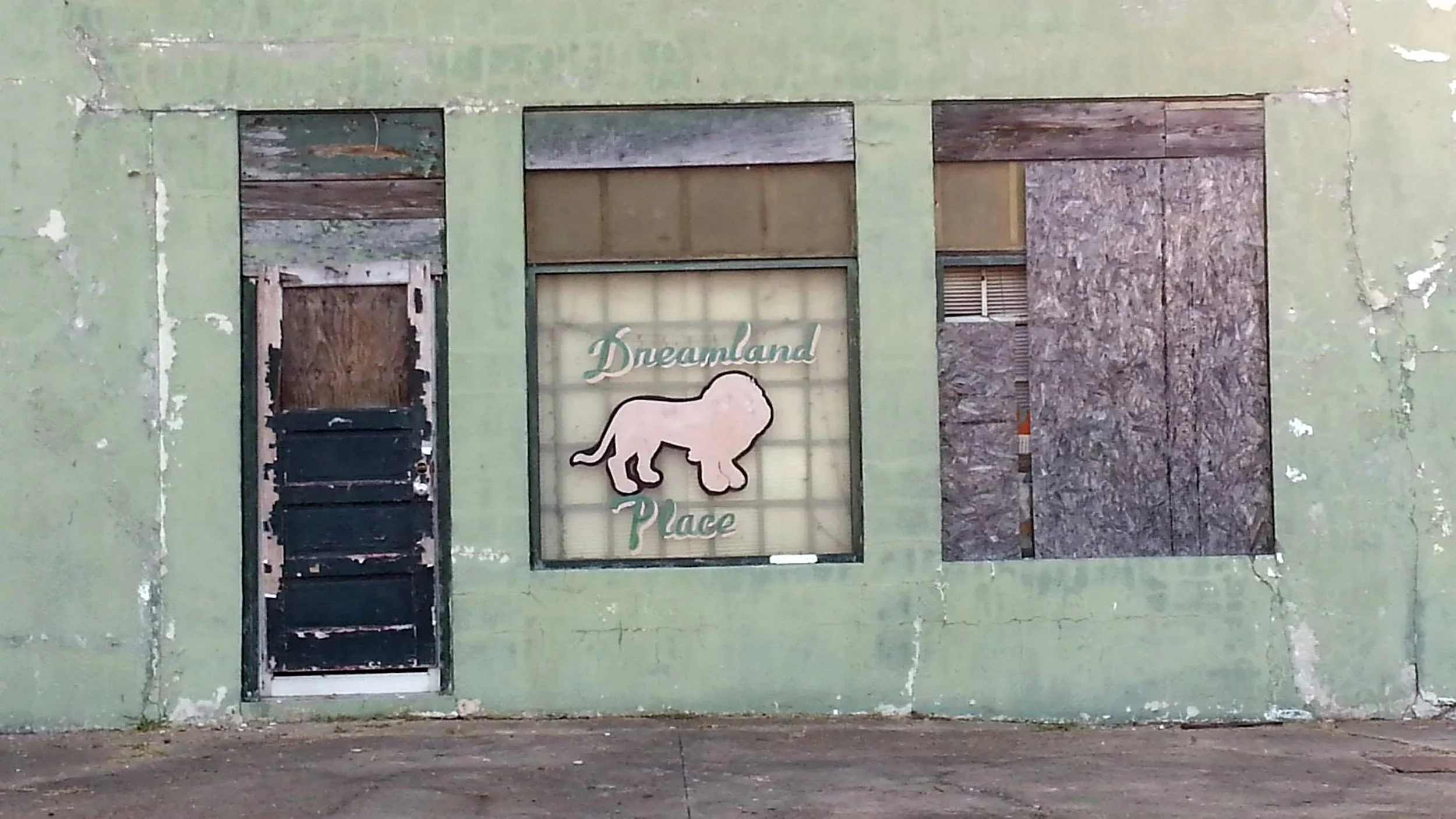 An old, weathered storefront with boarded-up windows and a green painted exterior wall. A sign reads "Dreamland Place" with a depiction of a lion.