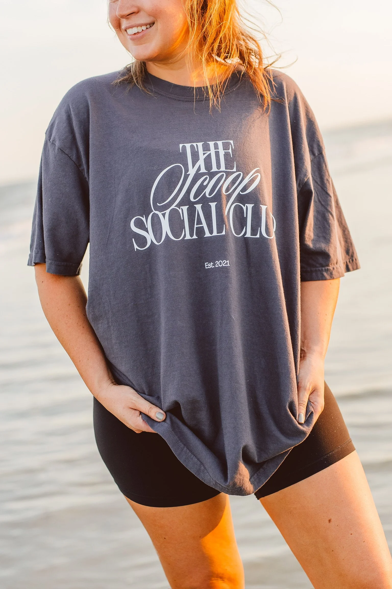 A person with red hair smiling while standing on a beach at sunset, wearing a gray t-shirt with the words 'The Scoop Social Club' and black shorts.
