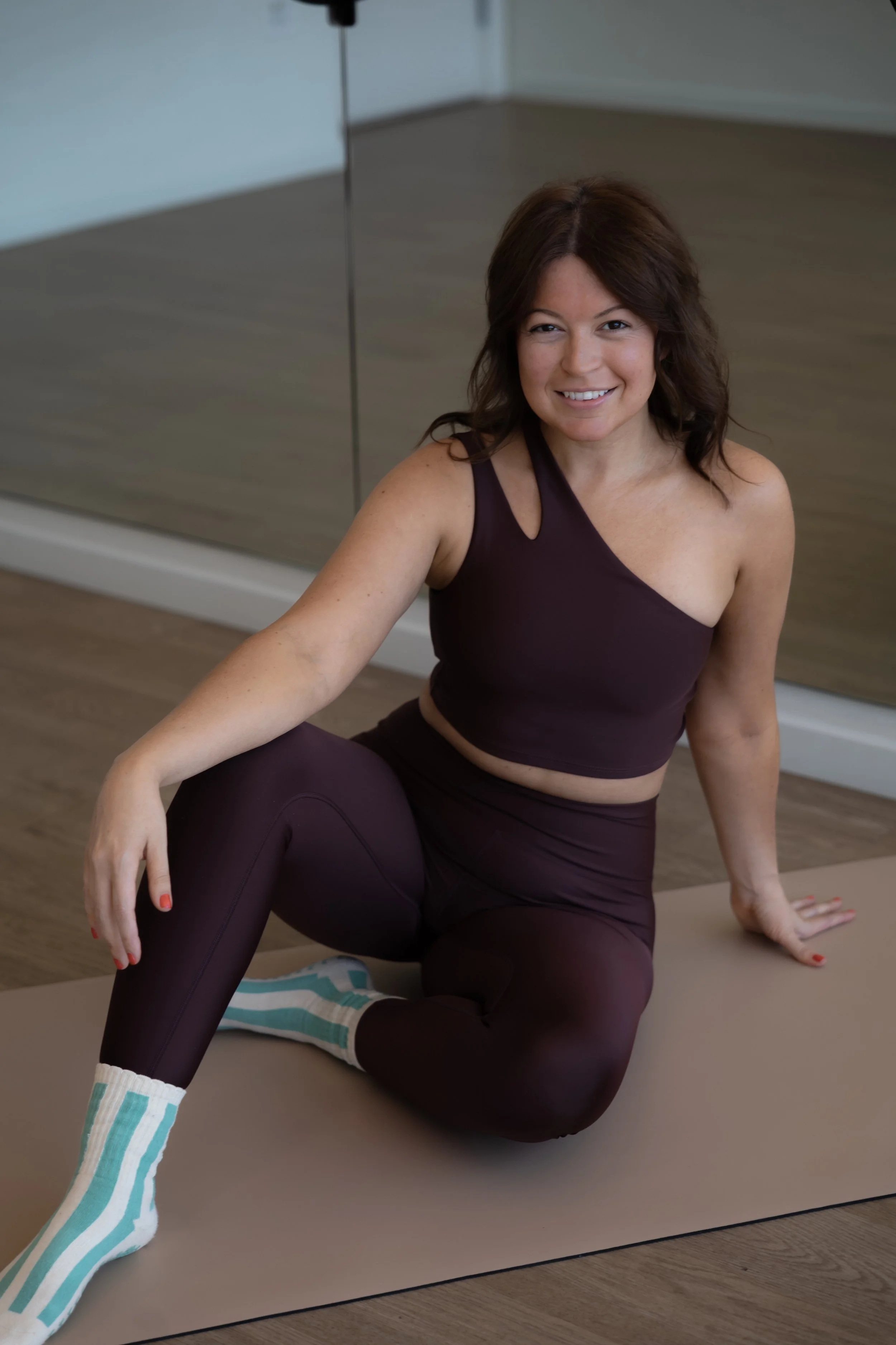 A woman smiling during a yoga or fitness session, sitting on a yoga mat in a room with wooden flooring and a mirrored wall, wearing a purple sports top, matching leggings, and striped socks.