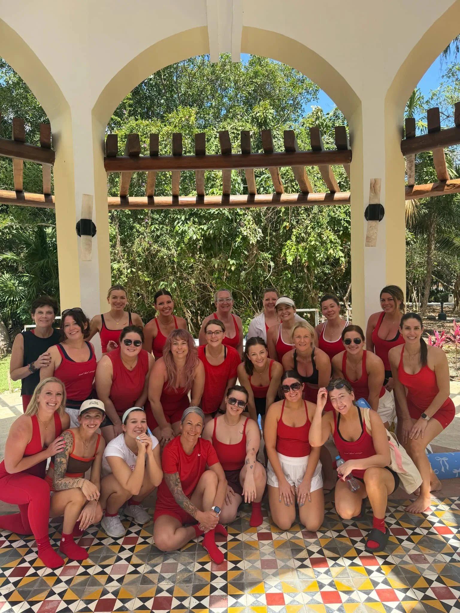 Group of women dressed in red and white casual clothing, smiling, in an outdoor setting with lush green trees and a tiled floor with colorful geometric patterns, under a white arch structure.