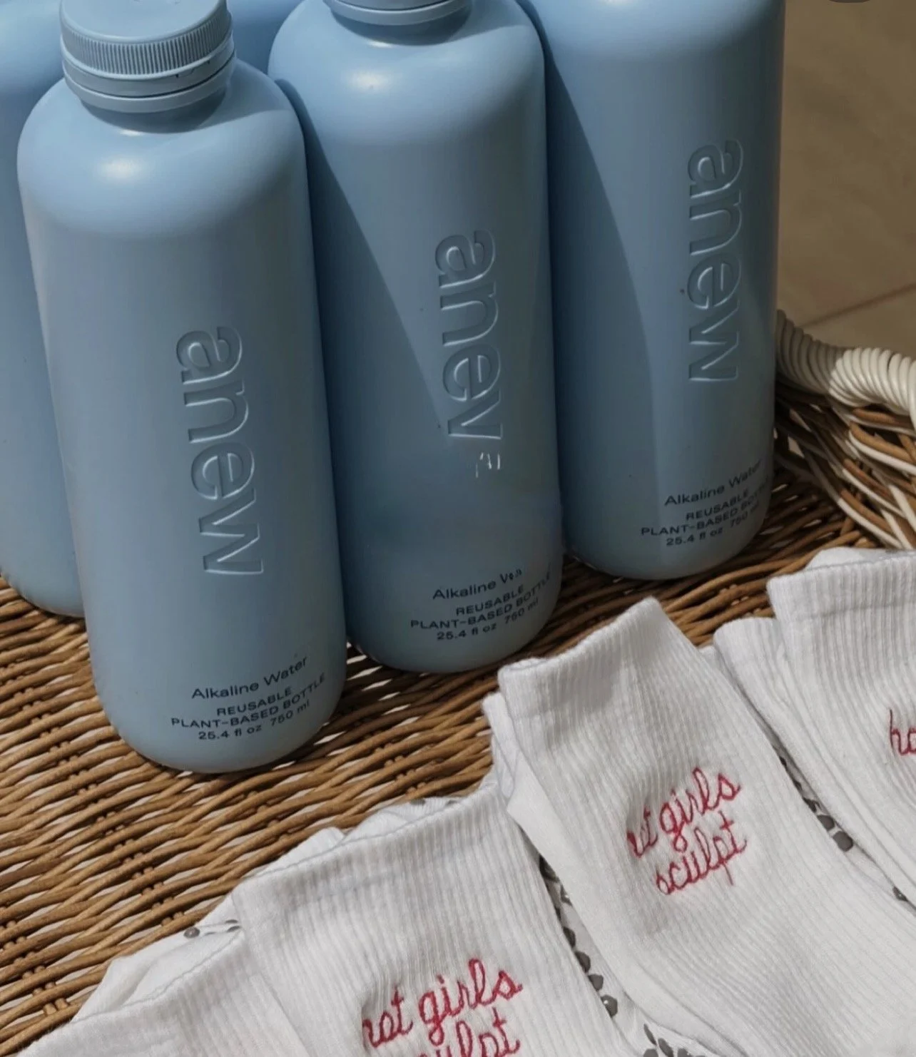 Three blue bottles labeled 'Alkaline Water' on a woven surface, with white cloths in front that have red embroidered text.