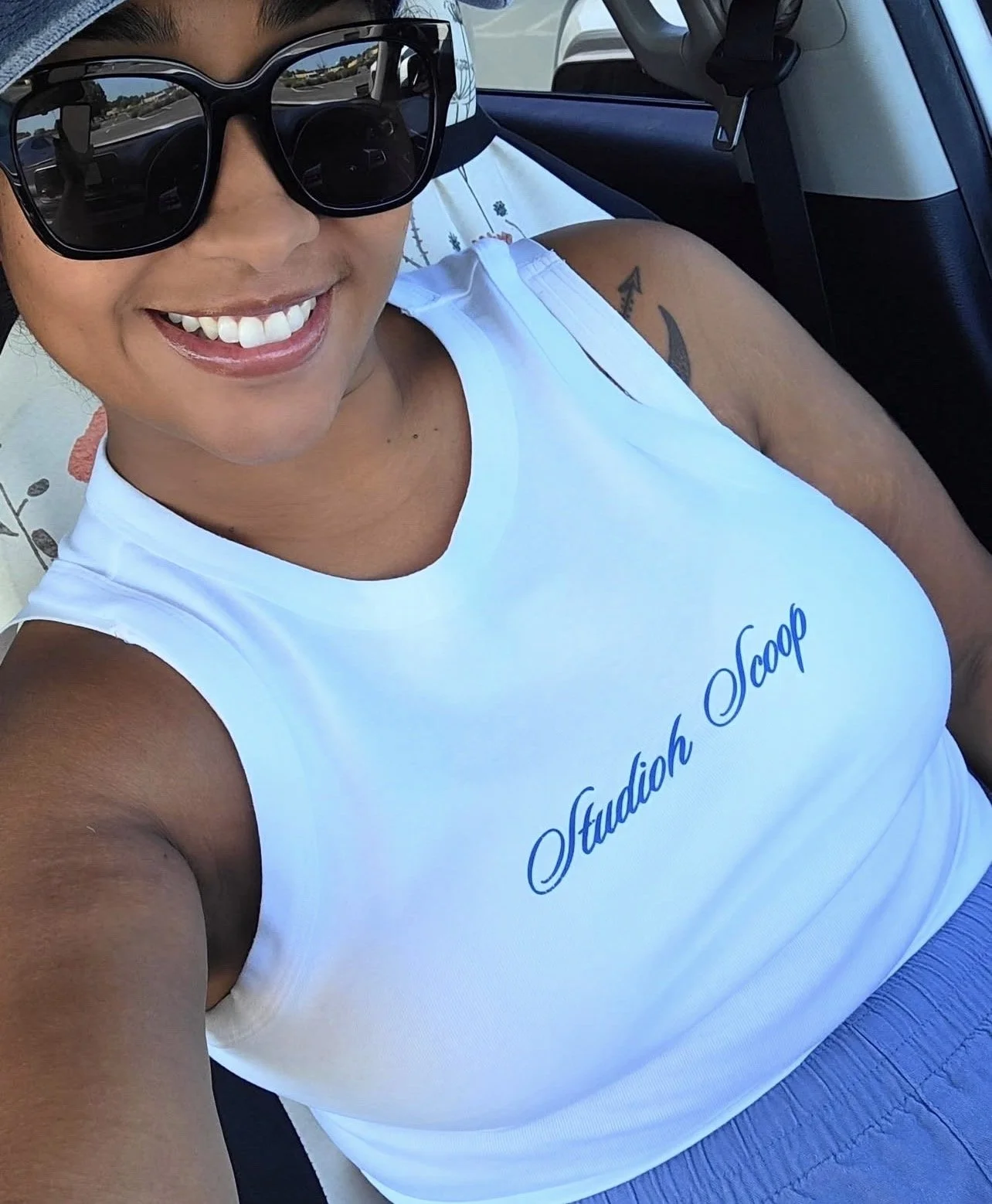 A woman smiling wearing large sunglasses and a white tank top with blue cursive text that reads "Studio Scoop", sitting in a vehicle.