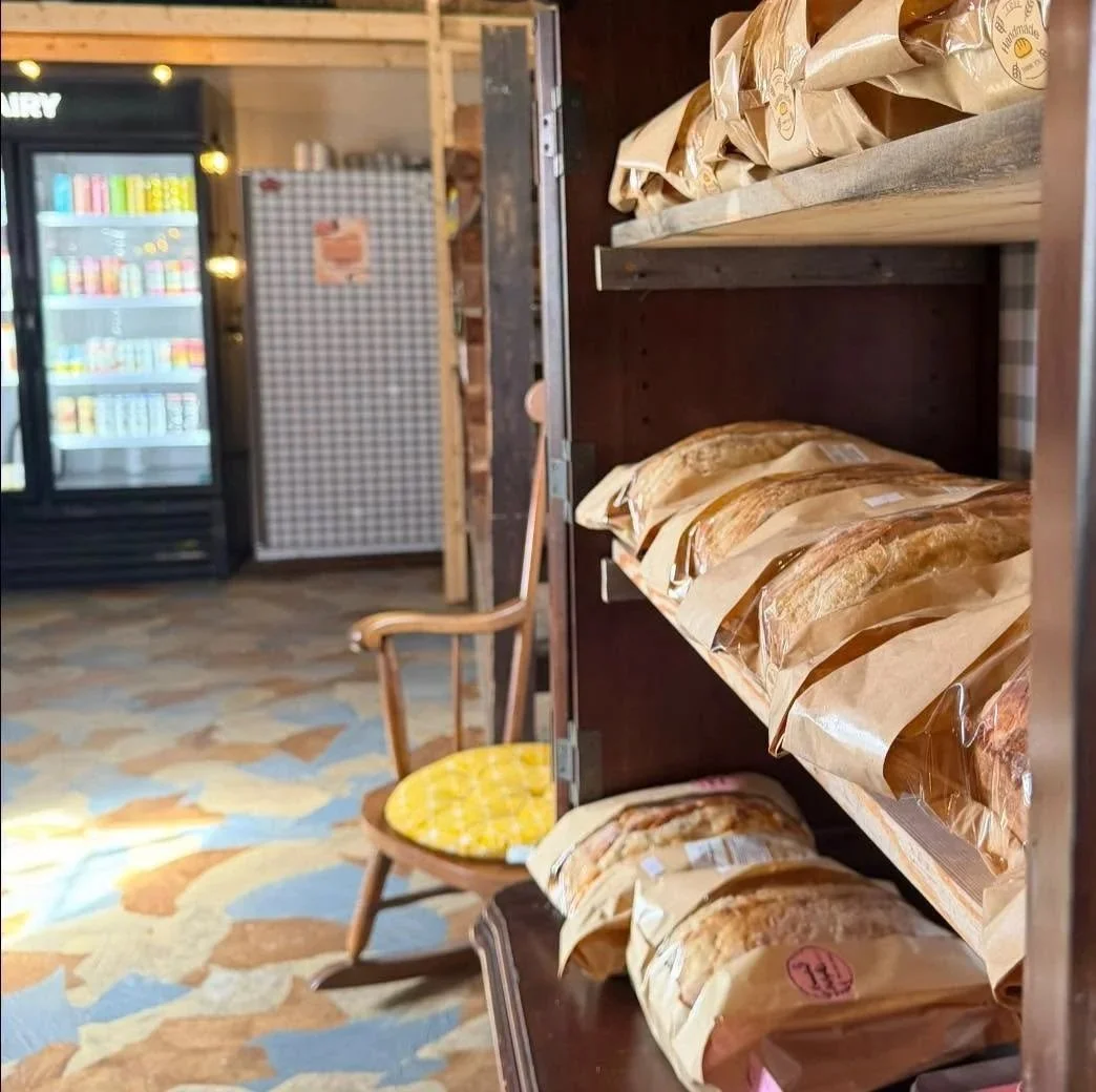 Shelf with multiple paper bags of baked goods or snacks in a store or bakery. A wooden chair with a yellow cushion is nearby, and a refrigerator or cold storage unit is in the background.