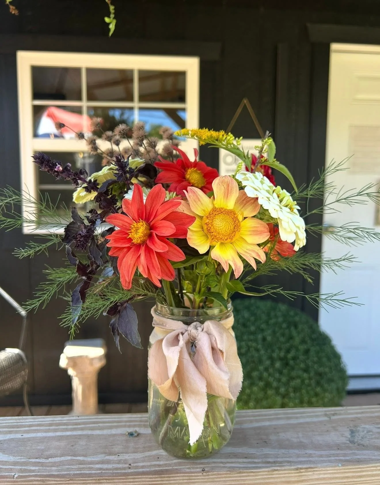 A bouquet of colorful flowers in a glass jar on a wooden surface, with a black background and a window reflecting the sky.