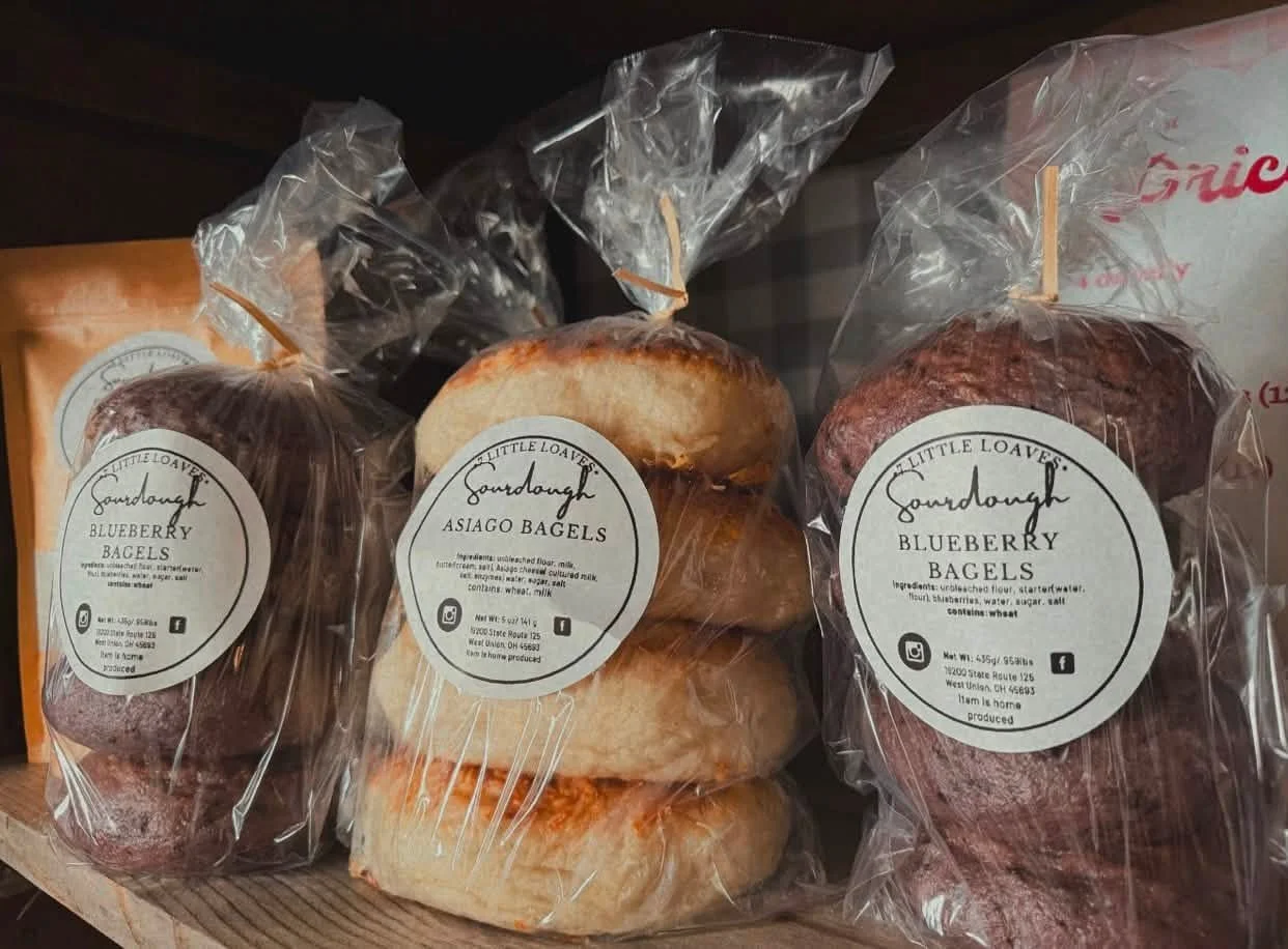 Three bags of bagels labeled blueberry and asiago, with a plain bagel in the middle on a wooden shelf.