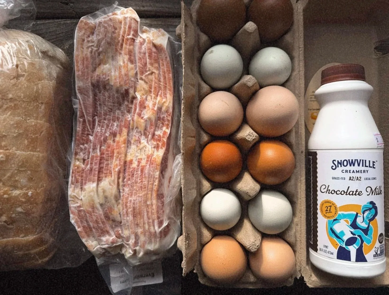 Fresh bacon, a carton of colorful eggs, a bottle of Snowville chocolate milk, and slices of bread on a dark surface.