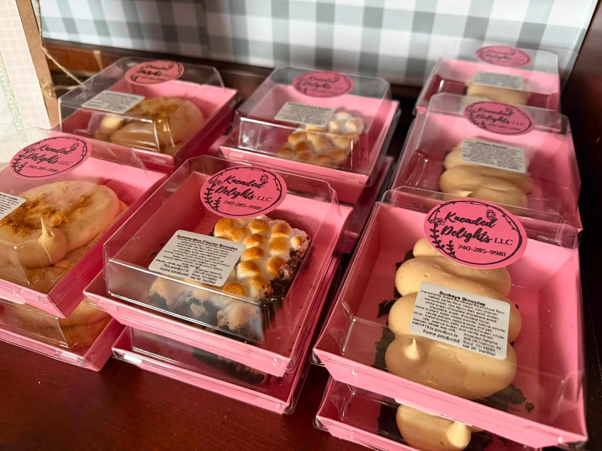 Several pink plastic boxes of baked goods are arranged on a dark red surface. Each box contains different baked items such as cookies, cupcakes, and other pastries. The boxes have a round pink sticker with black decorative writing that reads 'Kendall Delights LLC' and a phone number.