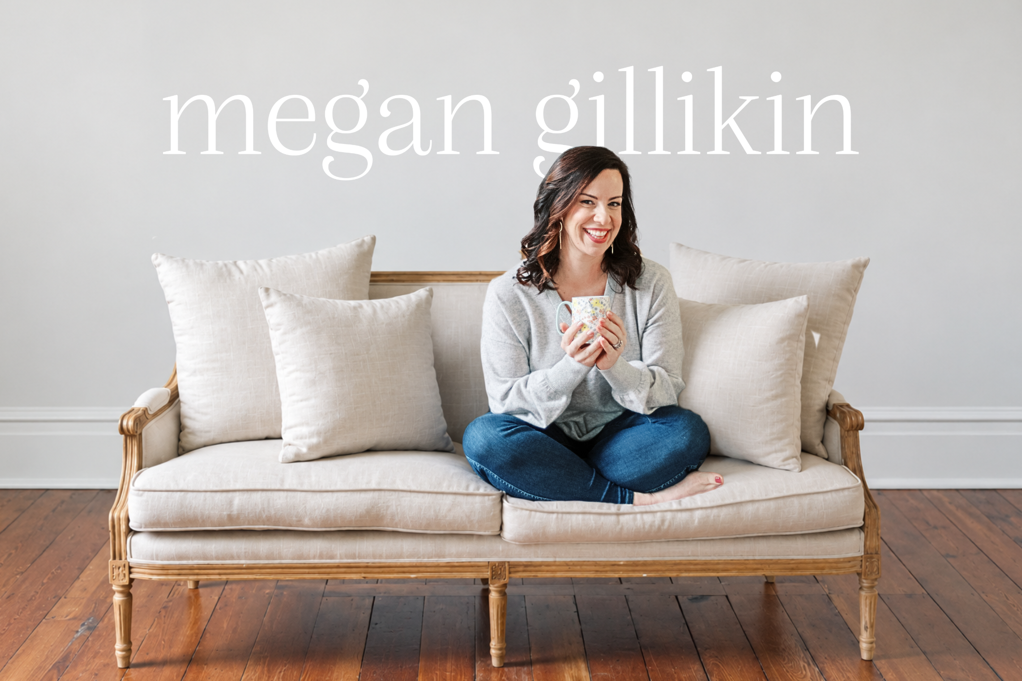 A woman sitting cross-legged on a beige couch with several pillows, smiling and holding a floral mug with both hands, in a room with wood flooring and a plain gray wall.
