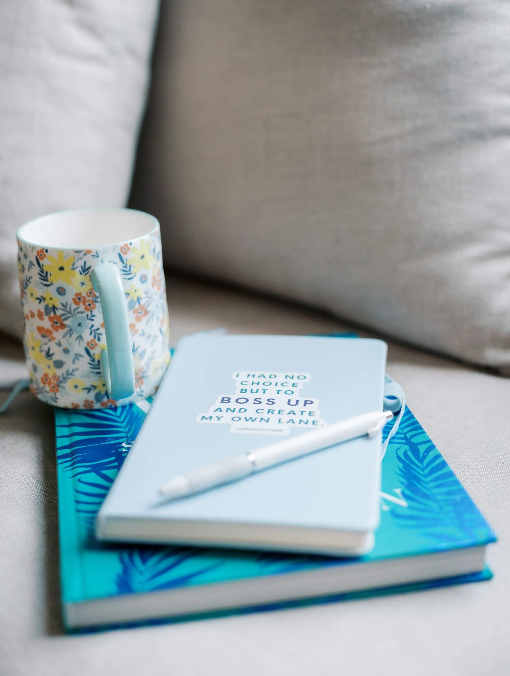 A floral mug, a notebook with a quote, a pen, and a blue folder with tropical leaf design resting on a beige sofa.