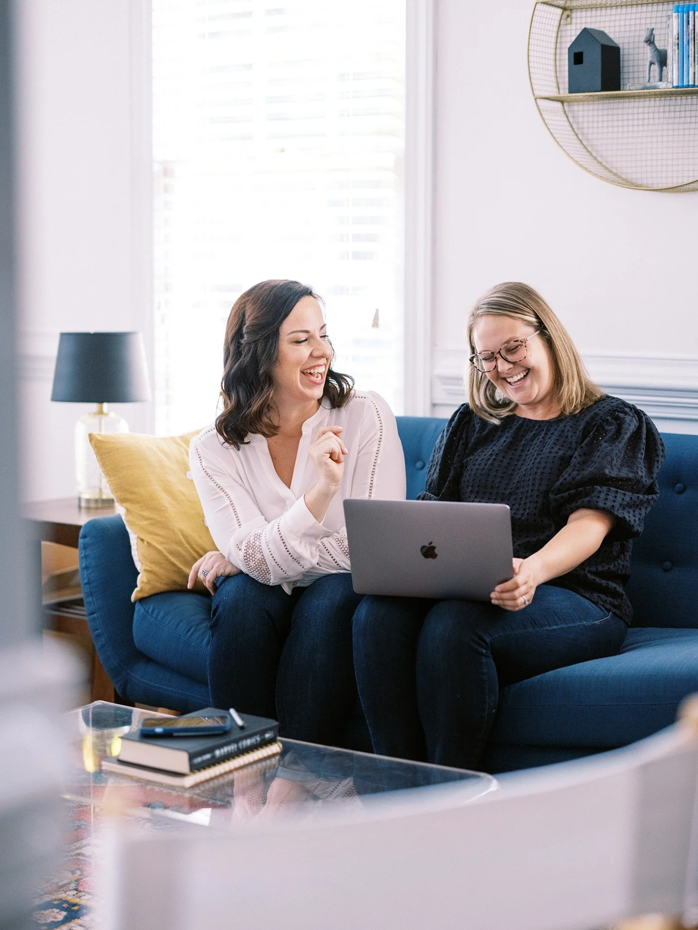 Two women sitting on a blue couch, looking at a laptop and laughing together in a bright living room.