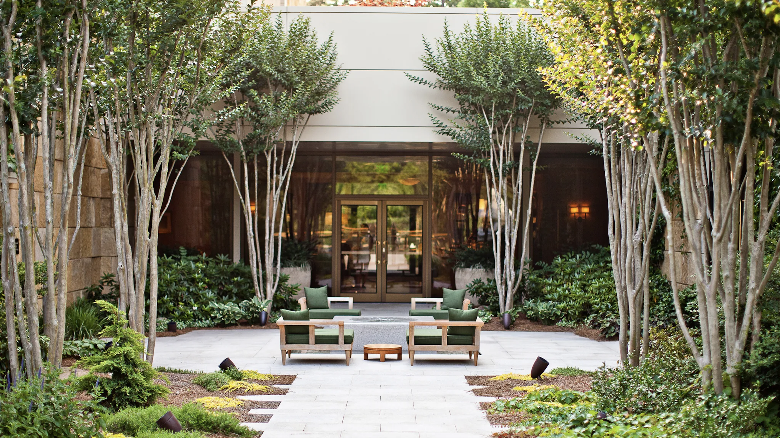 The Umstead Hotel outdoor patio with four green cushioned chairs and a small wooden table, surrounded by lush green trees and plants, leading to a building with glass doors.