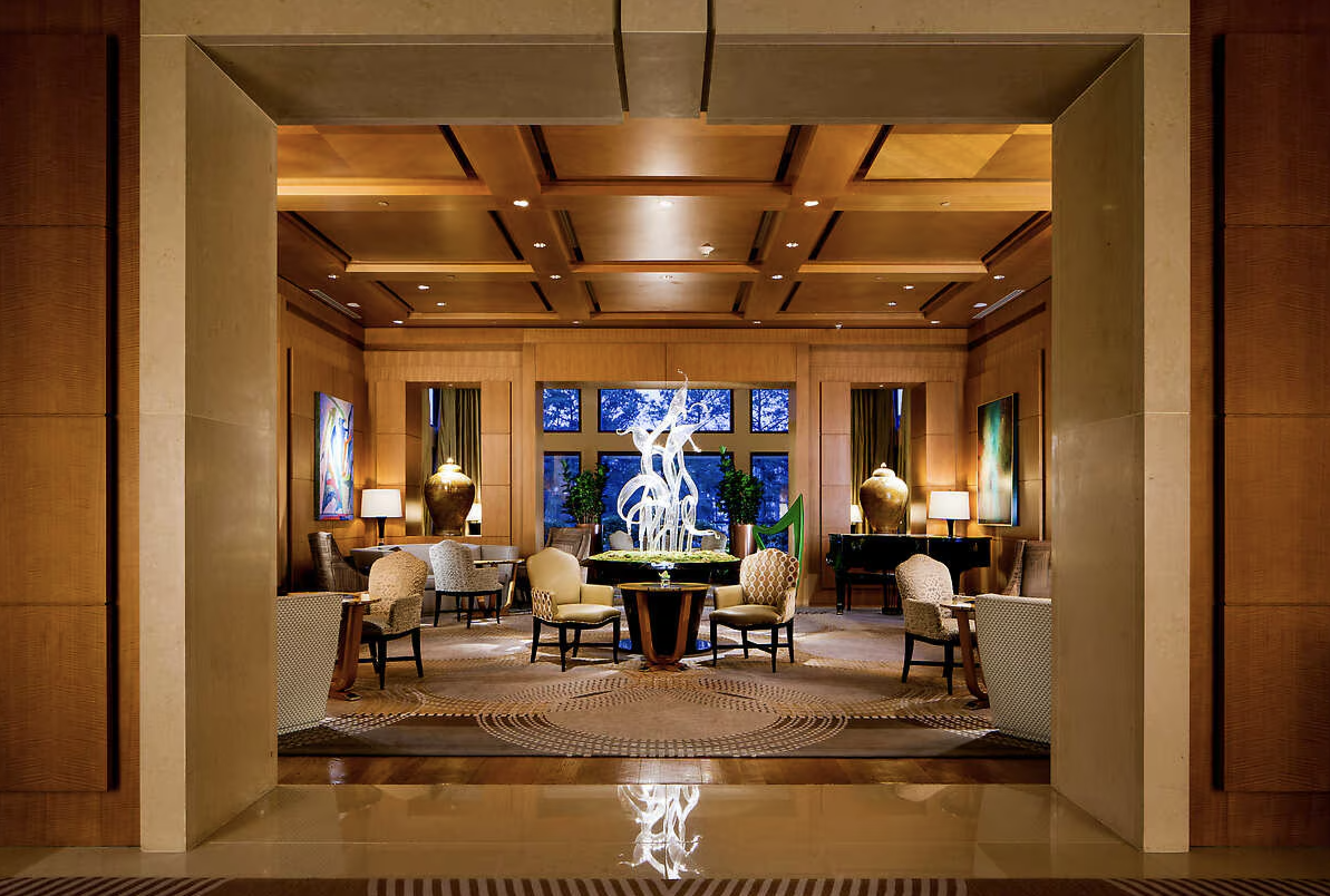 The Umsteadhotel lobby with wooden ceiling, seating area with armchairs and tables, large central sculpture, paintings on walls, lamps, and potted plants near windows showing trees outside.