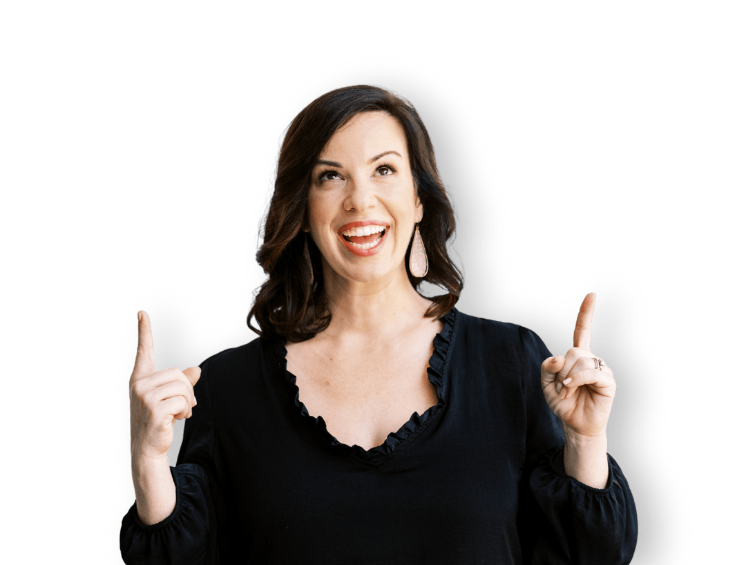 A woman with shoulder-length brown hair, wearing a black top and large earrings, smiling joyfully and pointing upward with both index fingers.