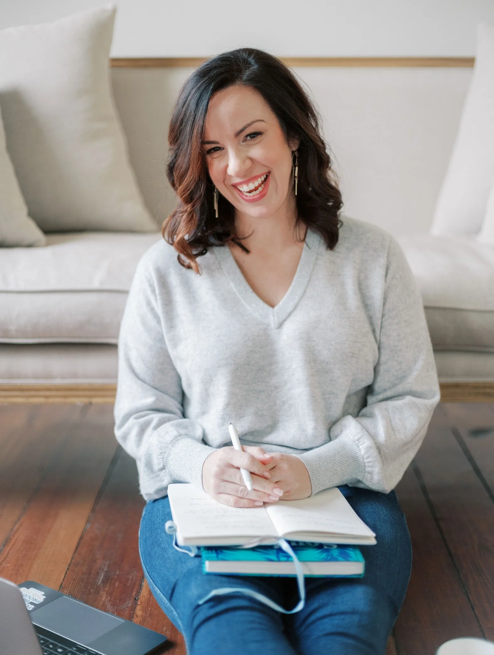 A woman with dark brown hair and earrings smiling, sitting on a wooden floor with a notebook on her lap, holding a pen, with a sofa and pillows in the background.