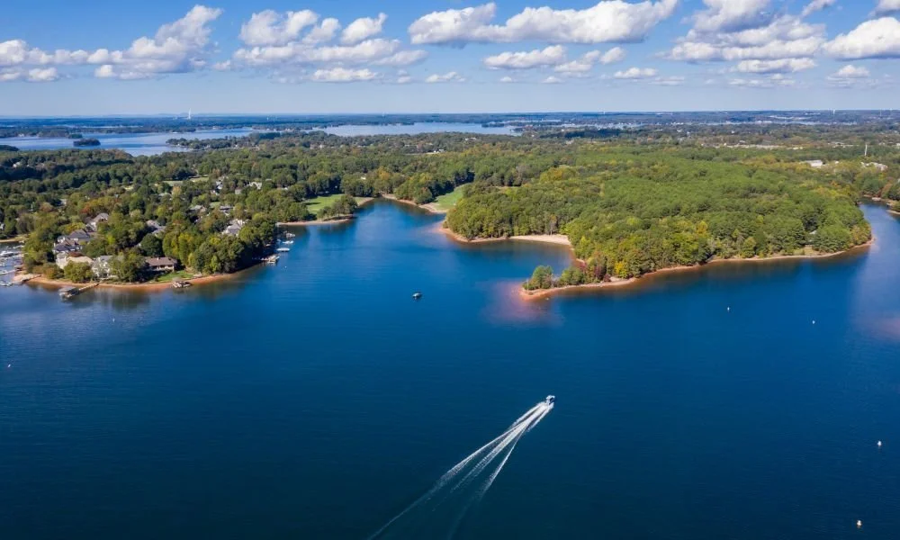Aerial view of a large, lush green island surrounded by a body of water, with boats and a boat jet stream visible on the water. The horizon shows a distant shoreline with more water and land.