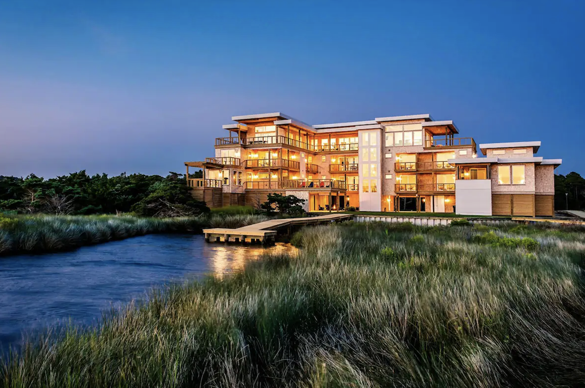 A modern multi-story house with large windows and balconies, lit up inside, situated near water with a grassy foreground.