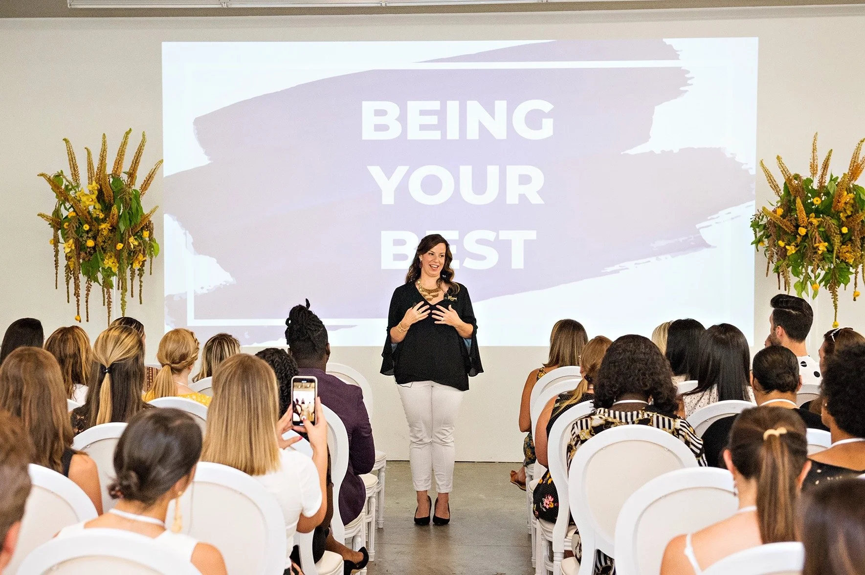 A woman standing and speaking in front of a seated audience during a presentation with the words "Being Your Best" displayed on a large screen behind her. The audience is diverse, and a person in the front row is capturing the moment with a cellphone. There are two large floral arrangements on either side of the stage.