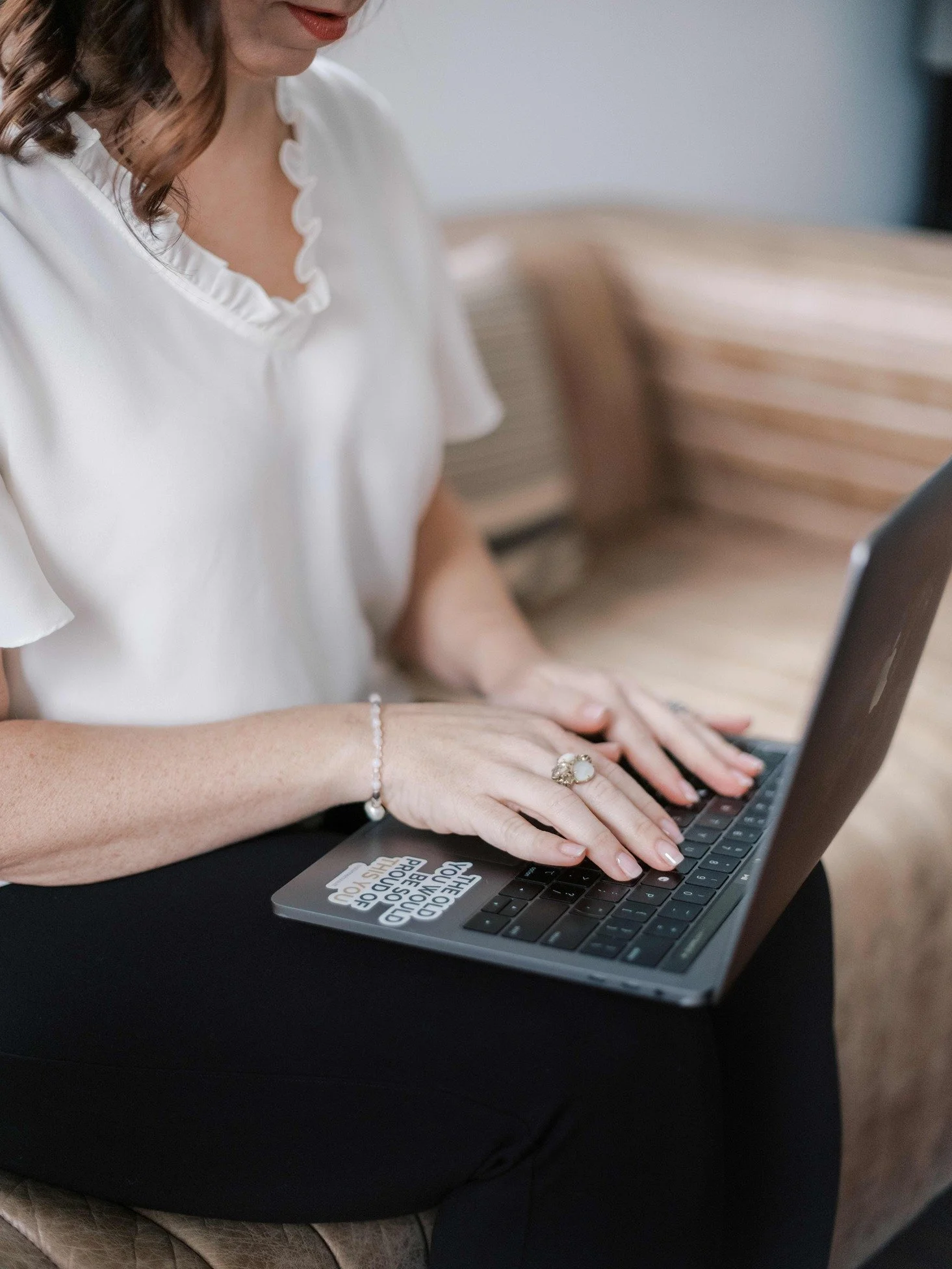 A woman wearing a white blouse with ruffled collar, typing on a laptop with a sticker that reads 'CERTIFIED I CAN PROVE I AM HAPPY.' She is seated on a cushioned surface, with a striped couch background.