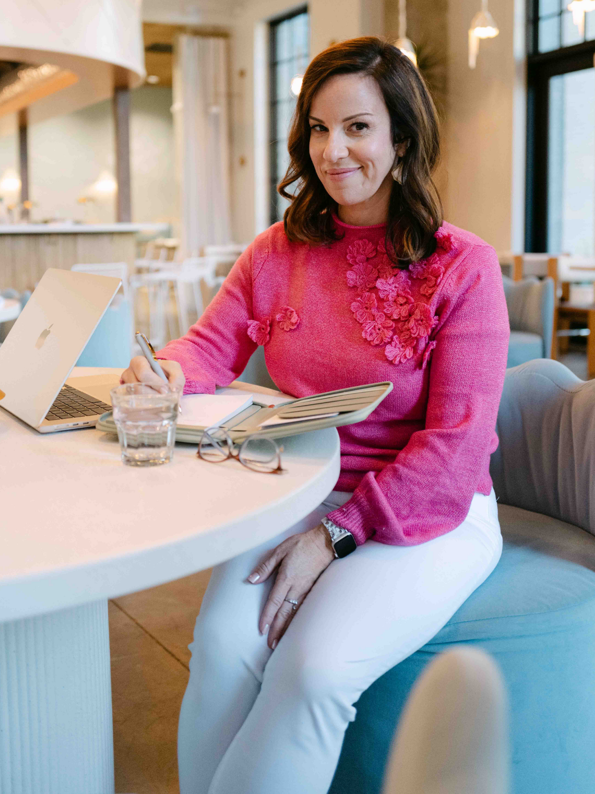 A woman with dark brown hair wearing a pink sweater with 3D floral embellishments on the upper chest sits at a white table in a bright, modern cafe. She is smiling and looking at the camera, with a notebook, pen, glasses, a glass of water, and a silver Apple laptop in front of her. There are large windows and warm lighting in the background.