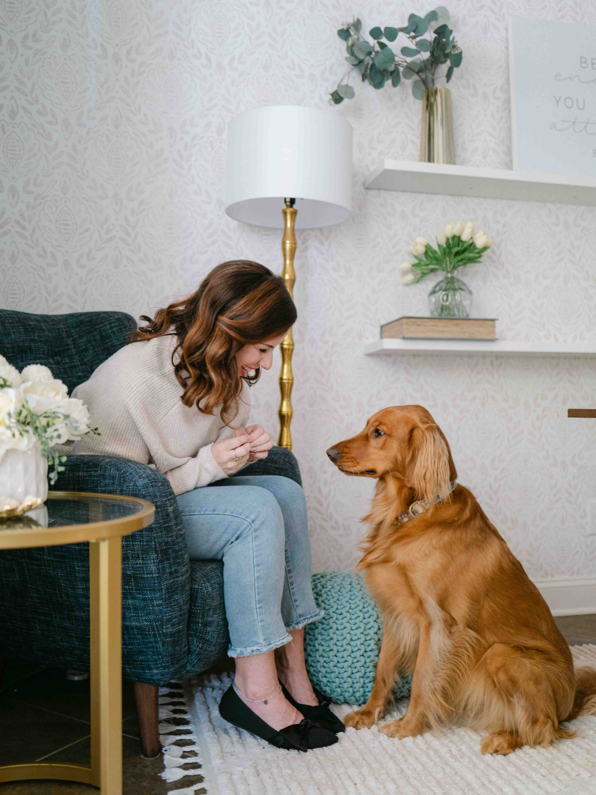 A woman sitting on a dark plaid armchair laughing and looking at a golden retriever dog sitting on the floor. The setting is a cozy living room with patterned wallpaper, a white lampshade on a gold stand, and shelves with vases and books.
