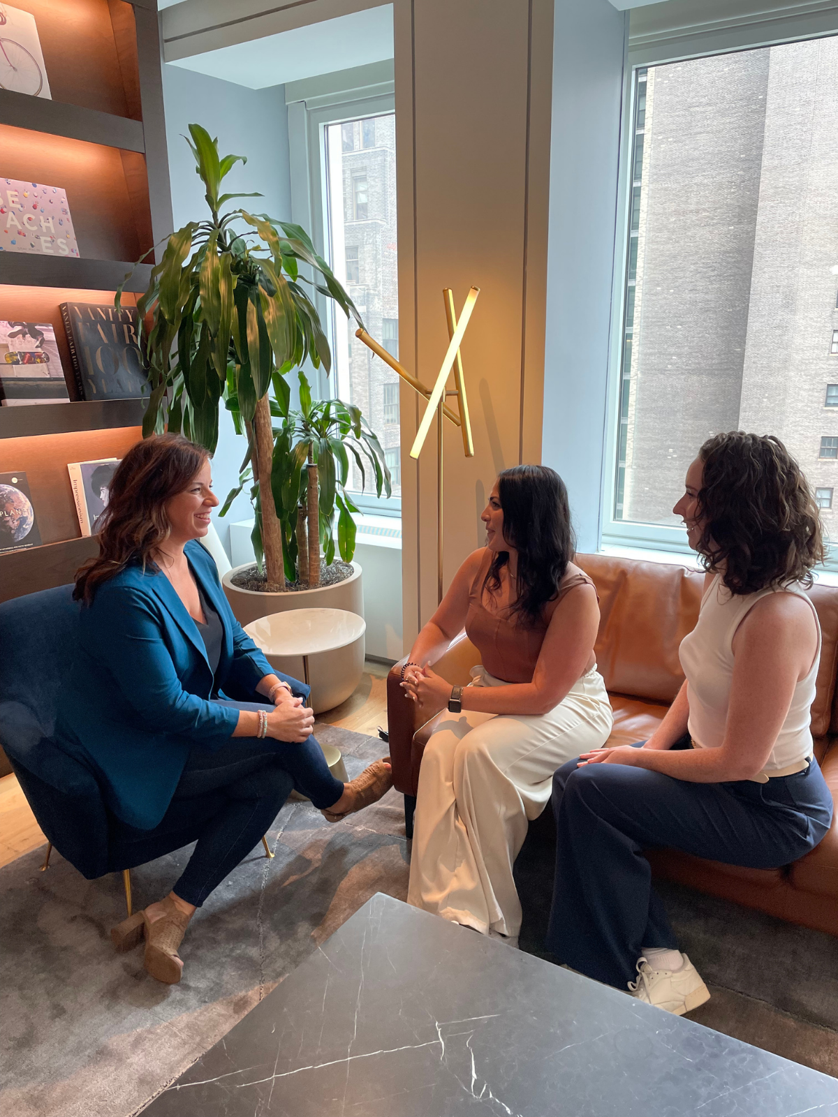 Three women sitting and talking in a modern office or lounge area, with large windows, a tall plant, and books on a shelf.