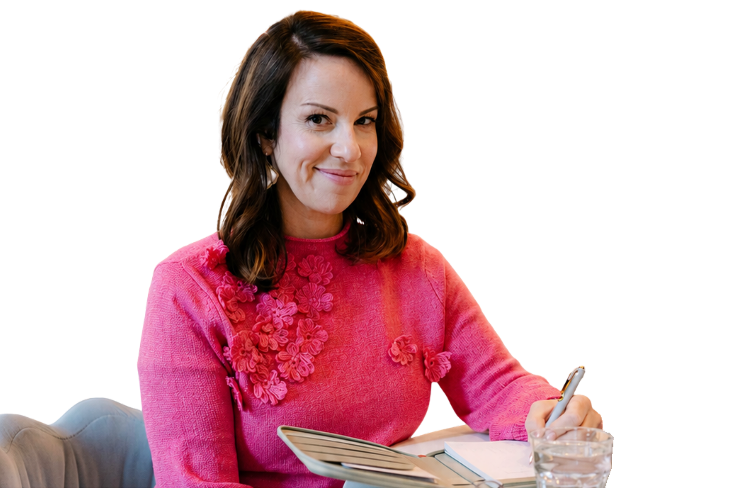 A woman with long brown hair, wearing a pink sweater with floral embellishments, is sitting at a table writing in a notebook with a pen. She is smiling slightly and looking directly at the camera. There is a glass of water and an open book on the table in front of her.