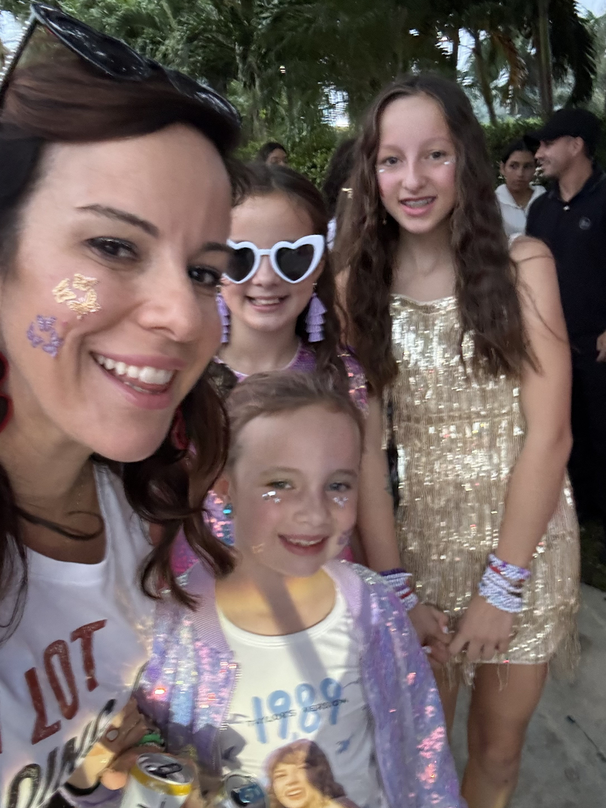 Group of women and girls at a festive outdoor event, dressed in sparkly outfits with face glitter, smiling and posing for a selfie.