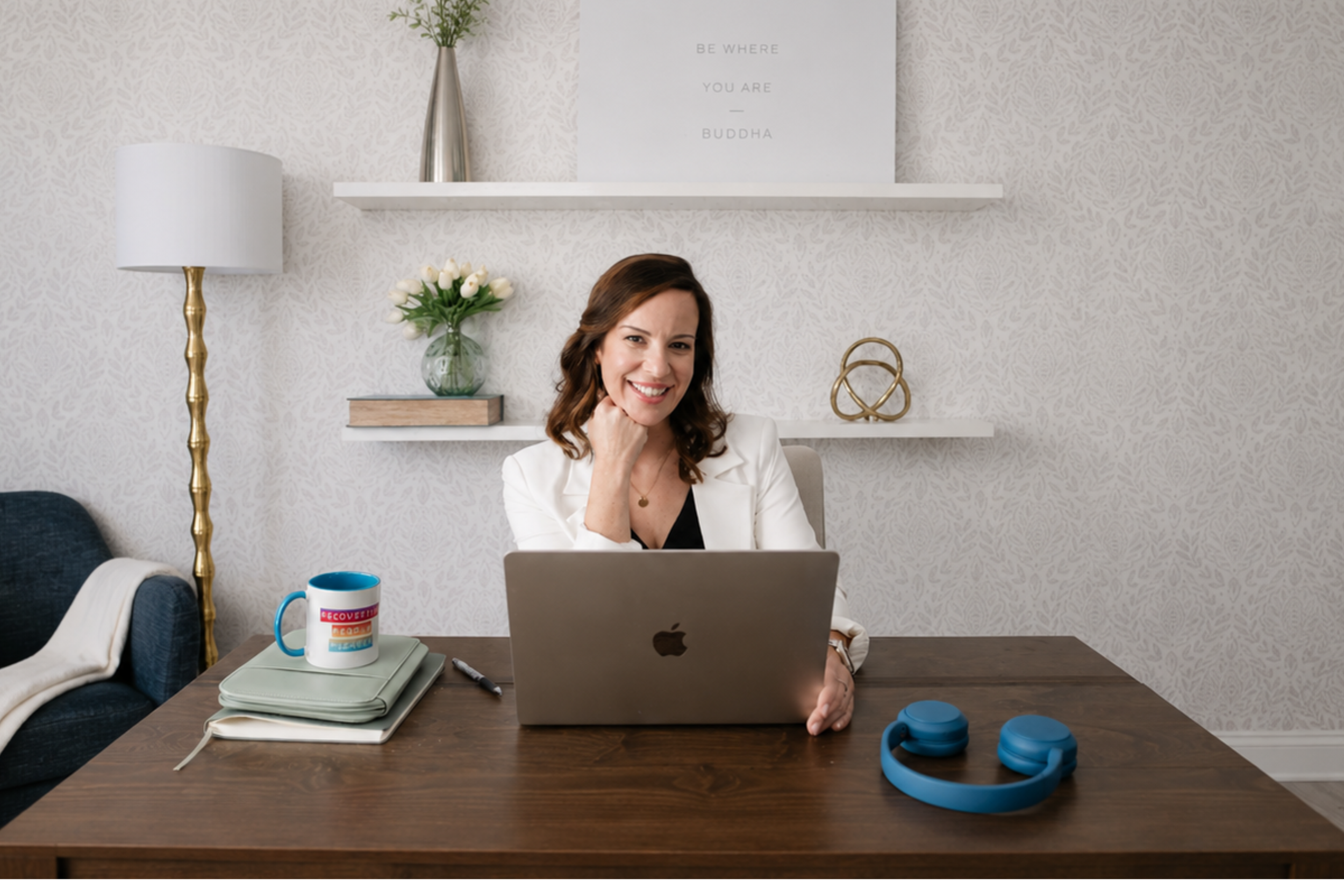 Woman with brown hair smiling at laptop in home office, with a white blazer and black top, surrounded by desk items including a mug, notebook, and blue headphones. Behind her are two white shelves with a green vase and white tulips, and a framed quote: 'Be where you are - Buddha.' A white lamp with a gold stand is on her left, and a blue couch with a white blanket is on her right.