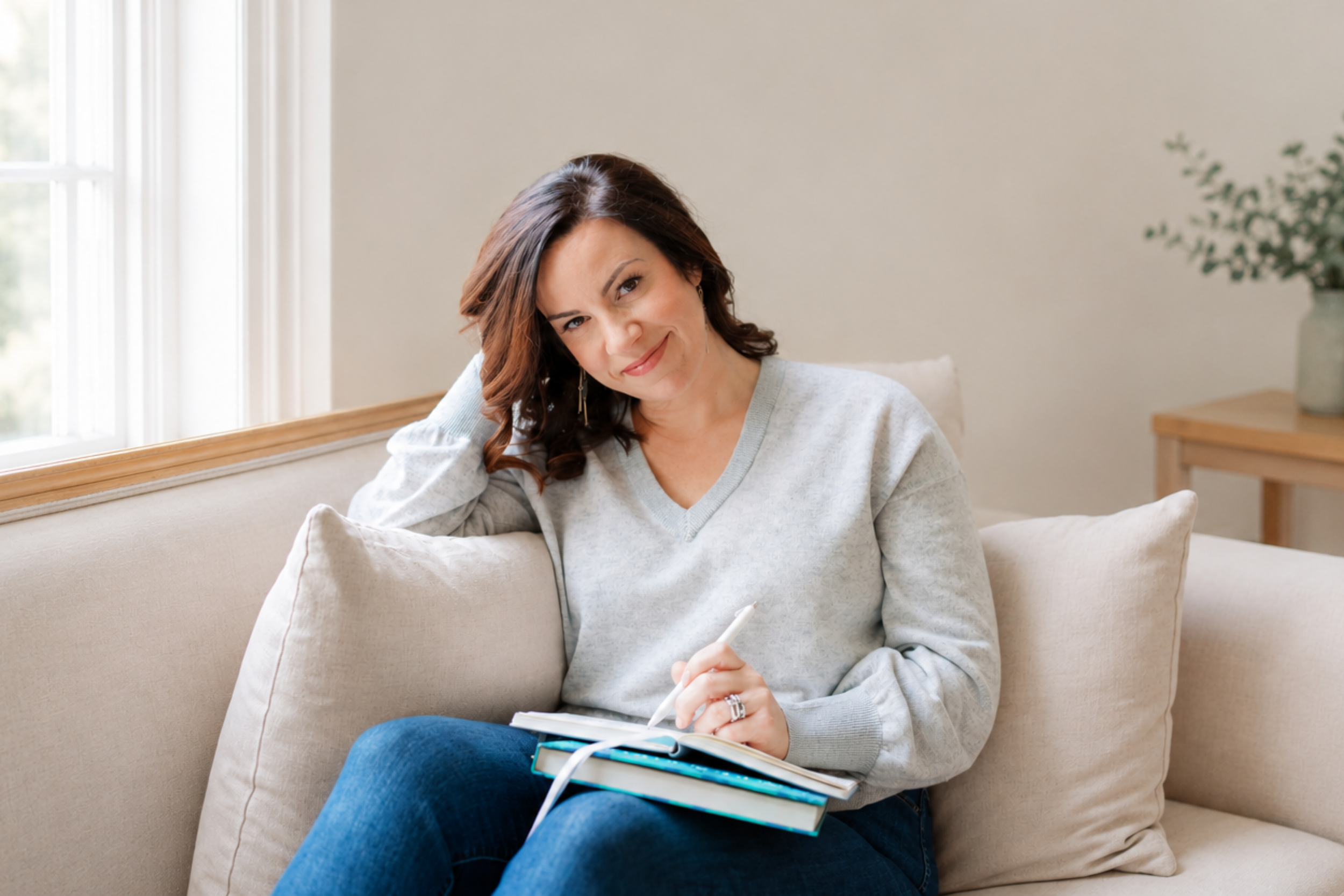 A woman with shoulder-length brown hair, wearing a light gray sweater, sitting on a beige couch, smiling and writing in a notebook with a pen. There is a window with white curtains behind her and a wooden table with a green potted plant to the right.