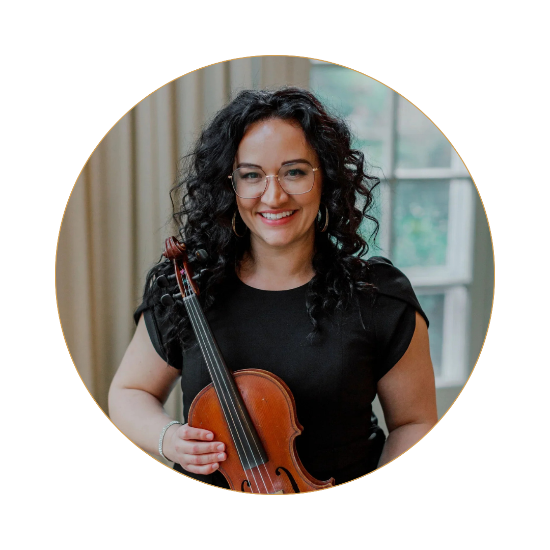 A smiling woman with curly black hair, glasses, and hoop earrings holding a violin inside a room with a window and beige curtains.