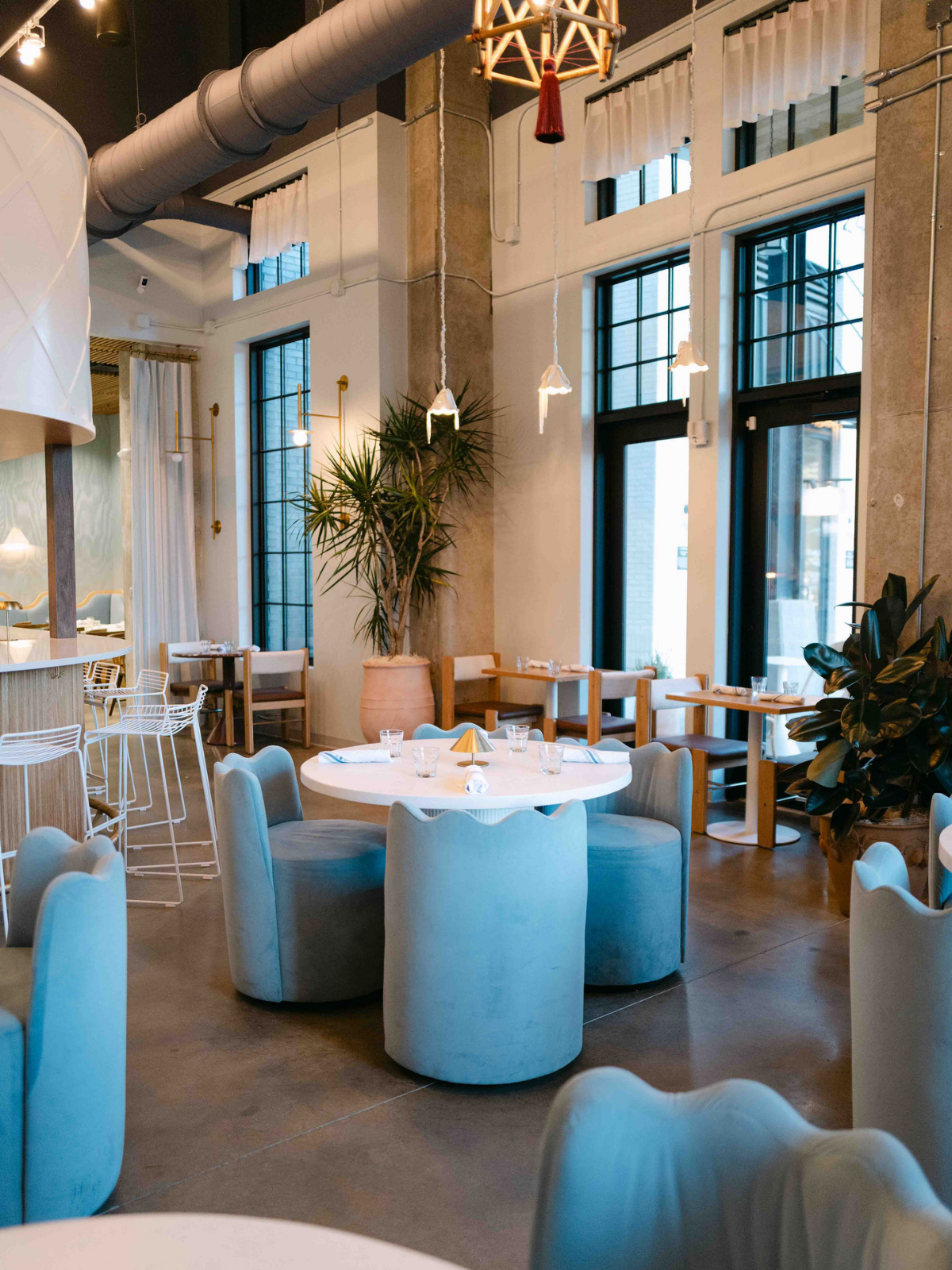 Interior of a modern restaurant with high ceilings, large windows, and contemporary decor. The space features a round table with light blue upholstered chairs, a potted plant, and elegant hanging lamps.
