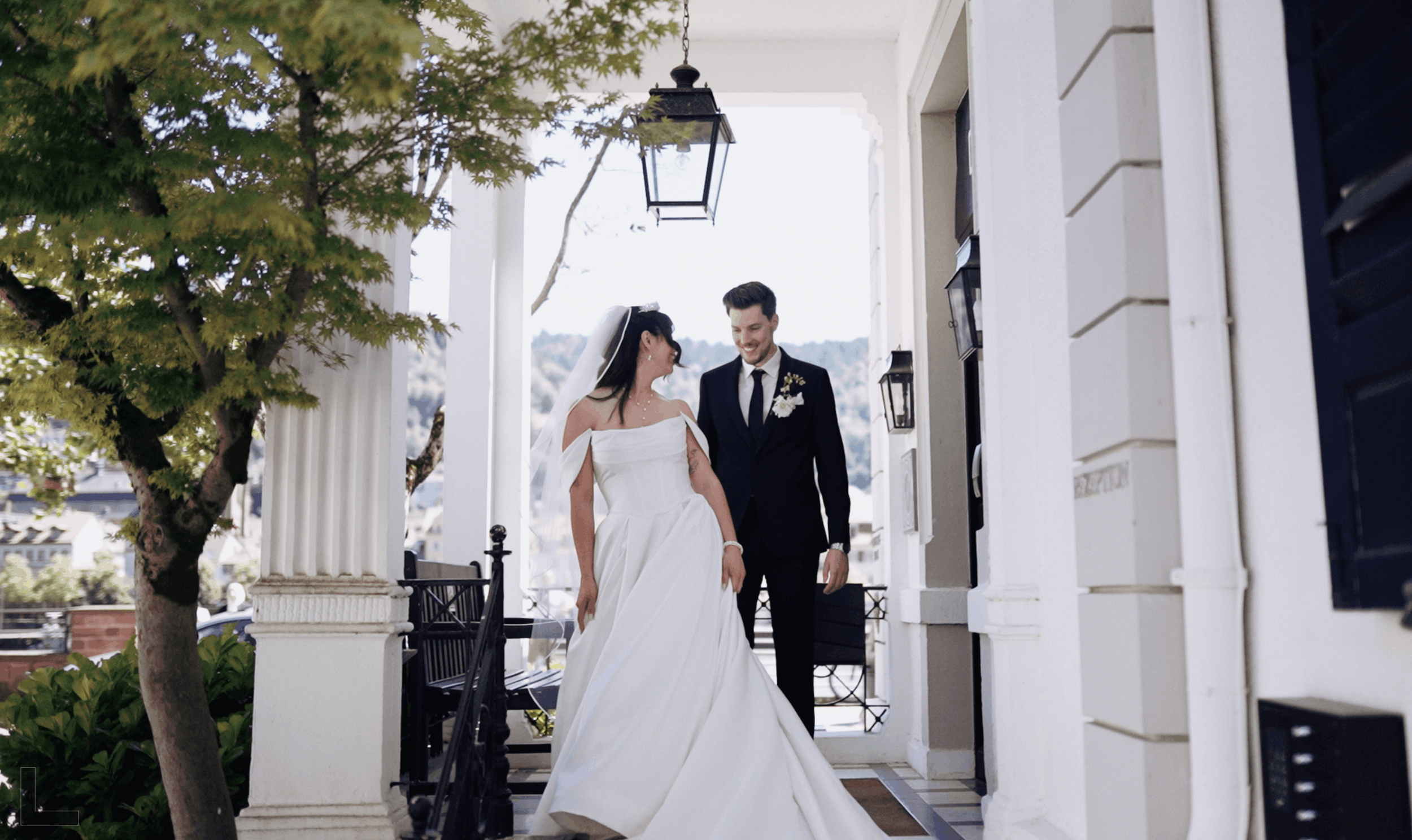 A bride and groom walking down the porch of a white house with columns, surrounded by greenery, with a tree on the left and a black lantern hanging from the ceiling.