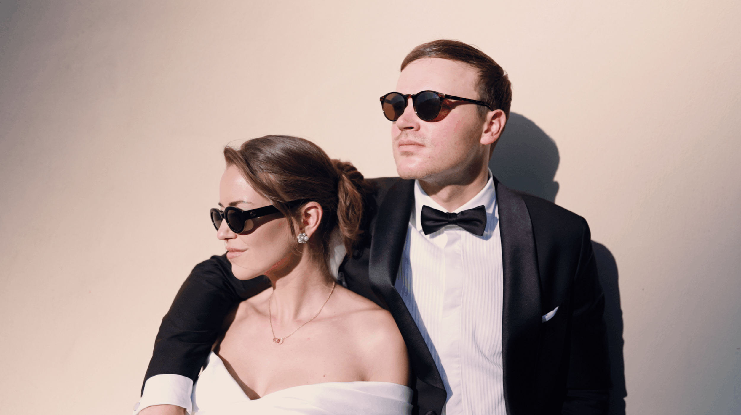 A man and woman wearing formal attire and sunglasses, standing close together against a plain background.