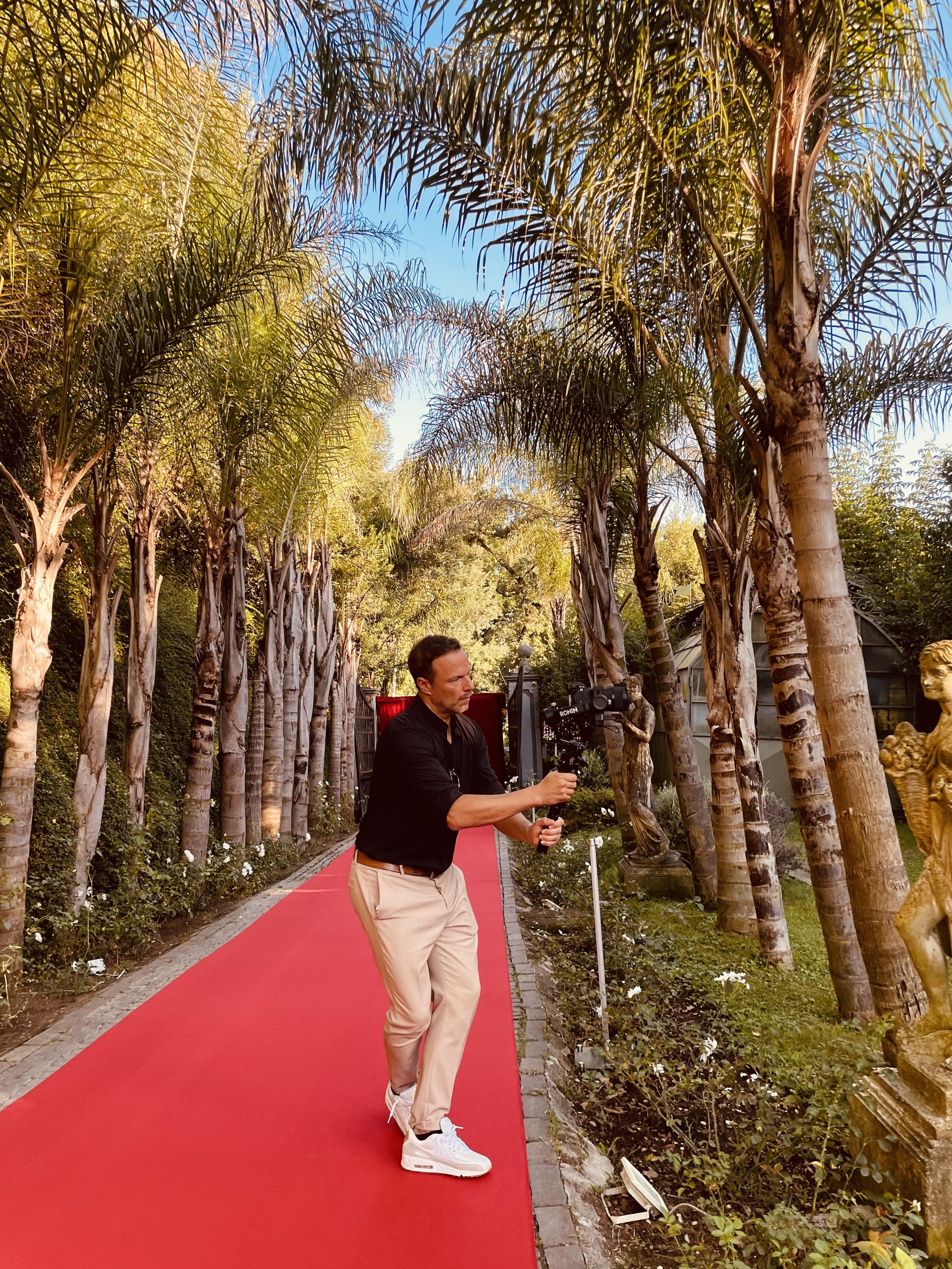 A man dressed in a black shirt, beige pants, and white sneakers is walking along a red carpet pathway lined with palm trees and lush greenery during daylight.