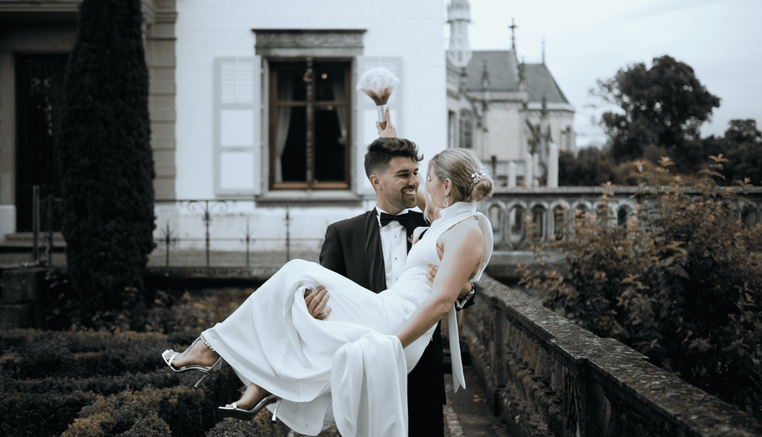 A bride and groom are outside in front of a white historic building, with the groom carrying the bride in his arms. The bride is holding a bouquet above her head, and both are smiling.