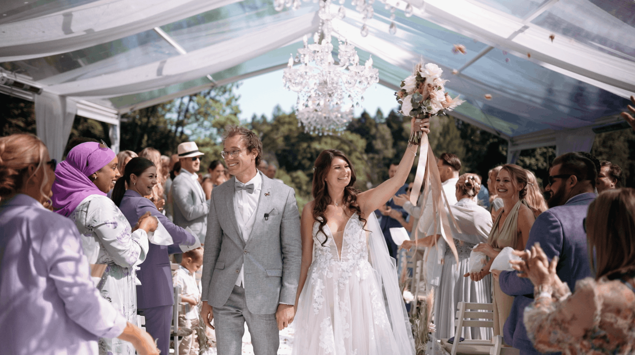 A bride and groom walking hand in hand through a reception decorated with a chandelier and floral arrangements, surrounded by guests in a bright outdoor setting.