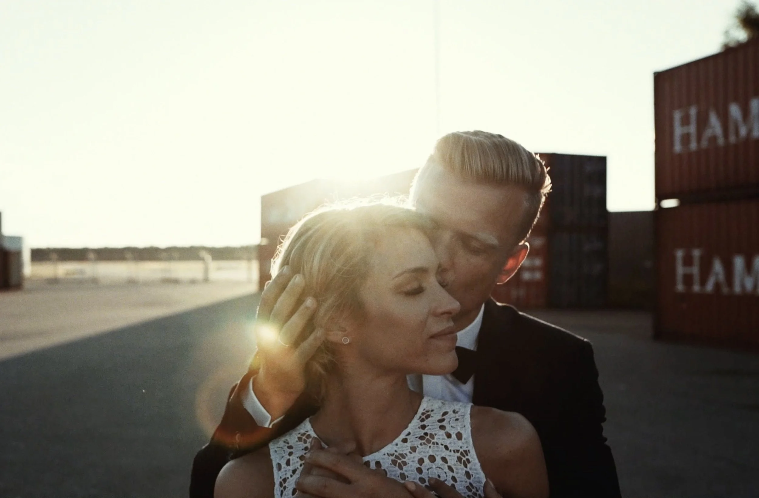 A couple embracing and kissing outdoors during sunset, with shipping containers and a highway in the background.