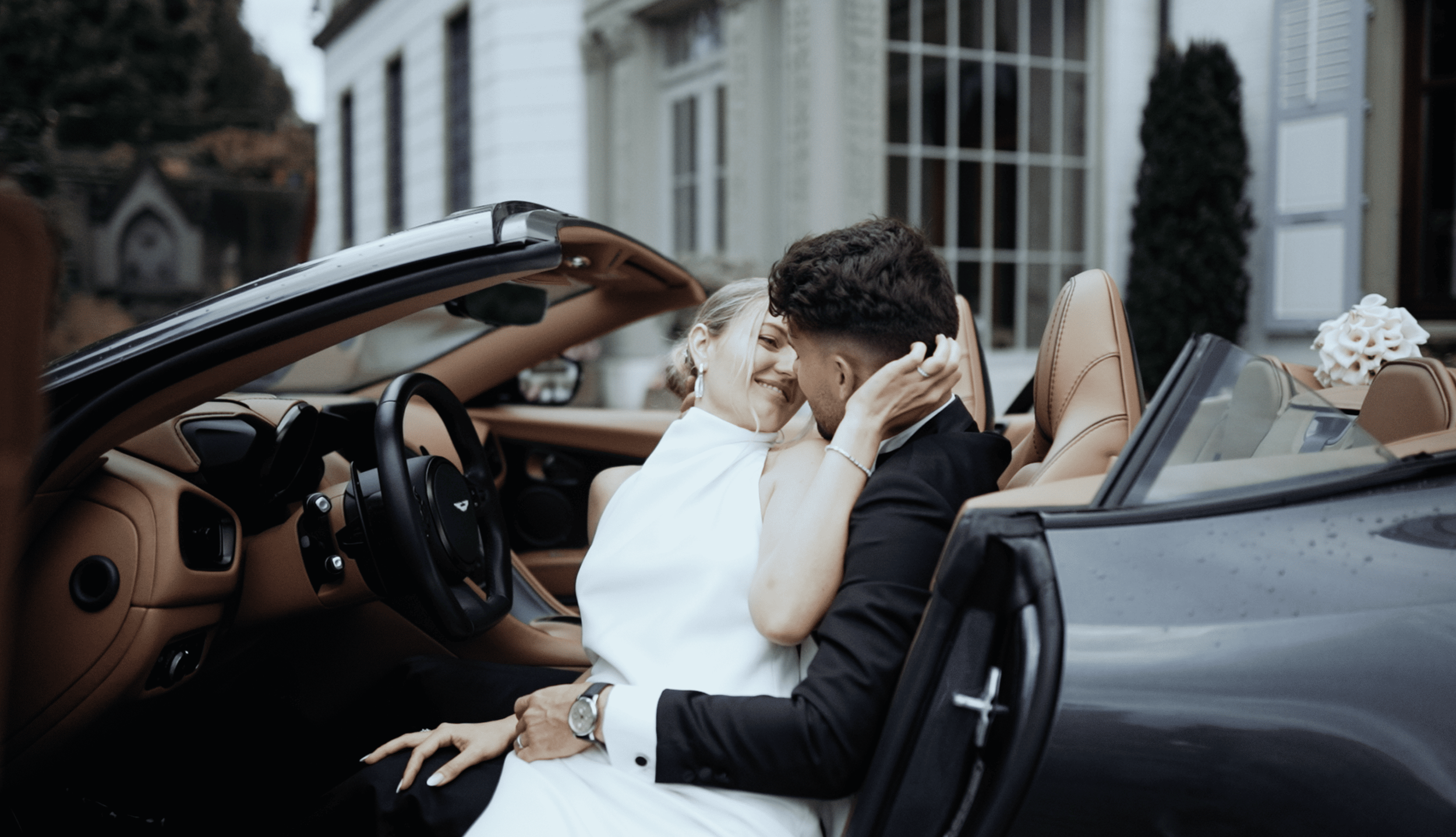 A couple in wedding attire sitting in a convertible car, smiling and embracing each other, outside a house.