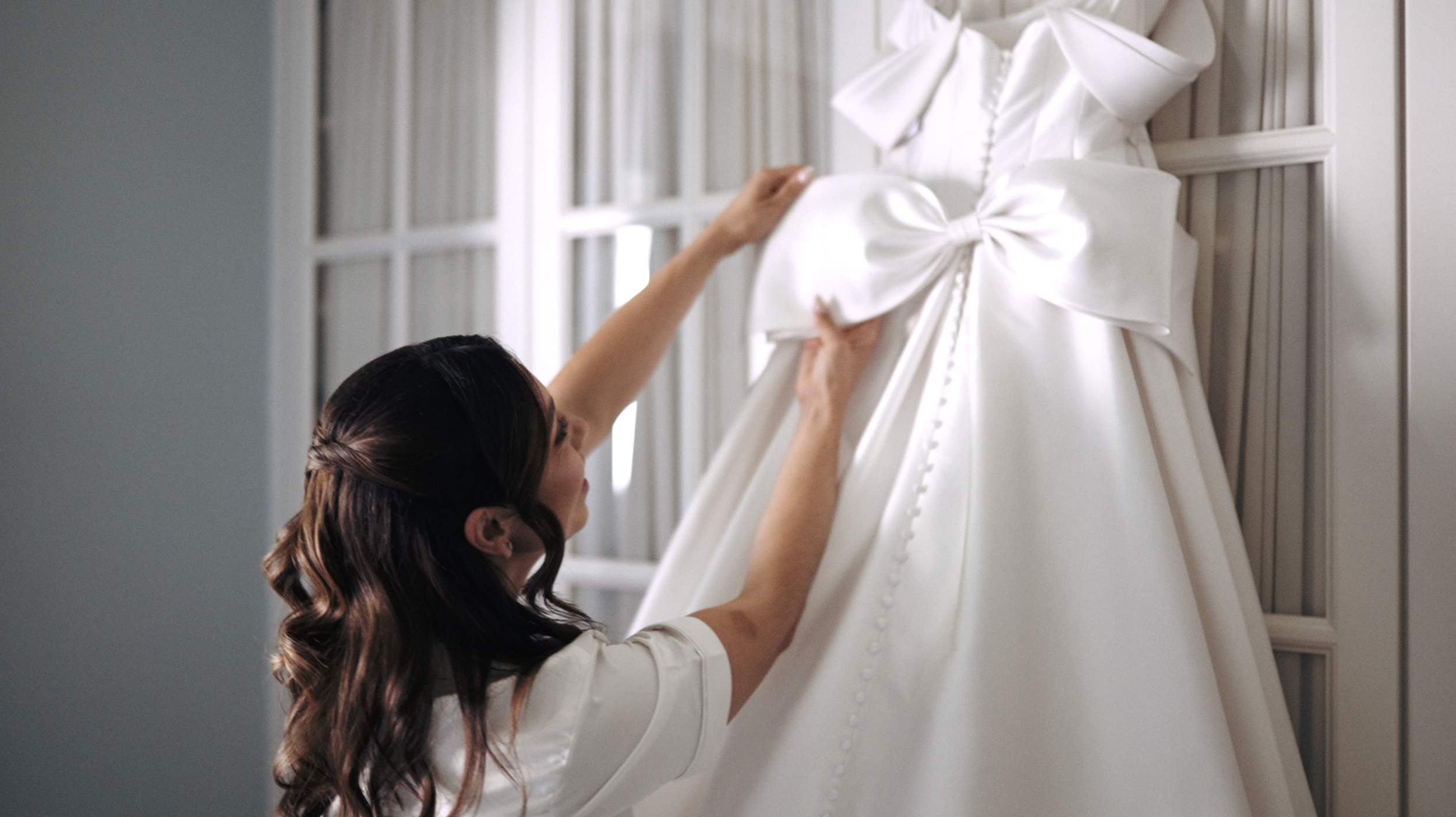Woman reaching to adjust a white wedding dress with a large bow, hanging on a door or wall.