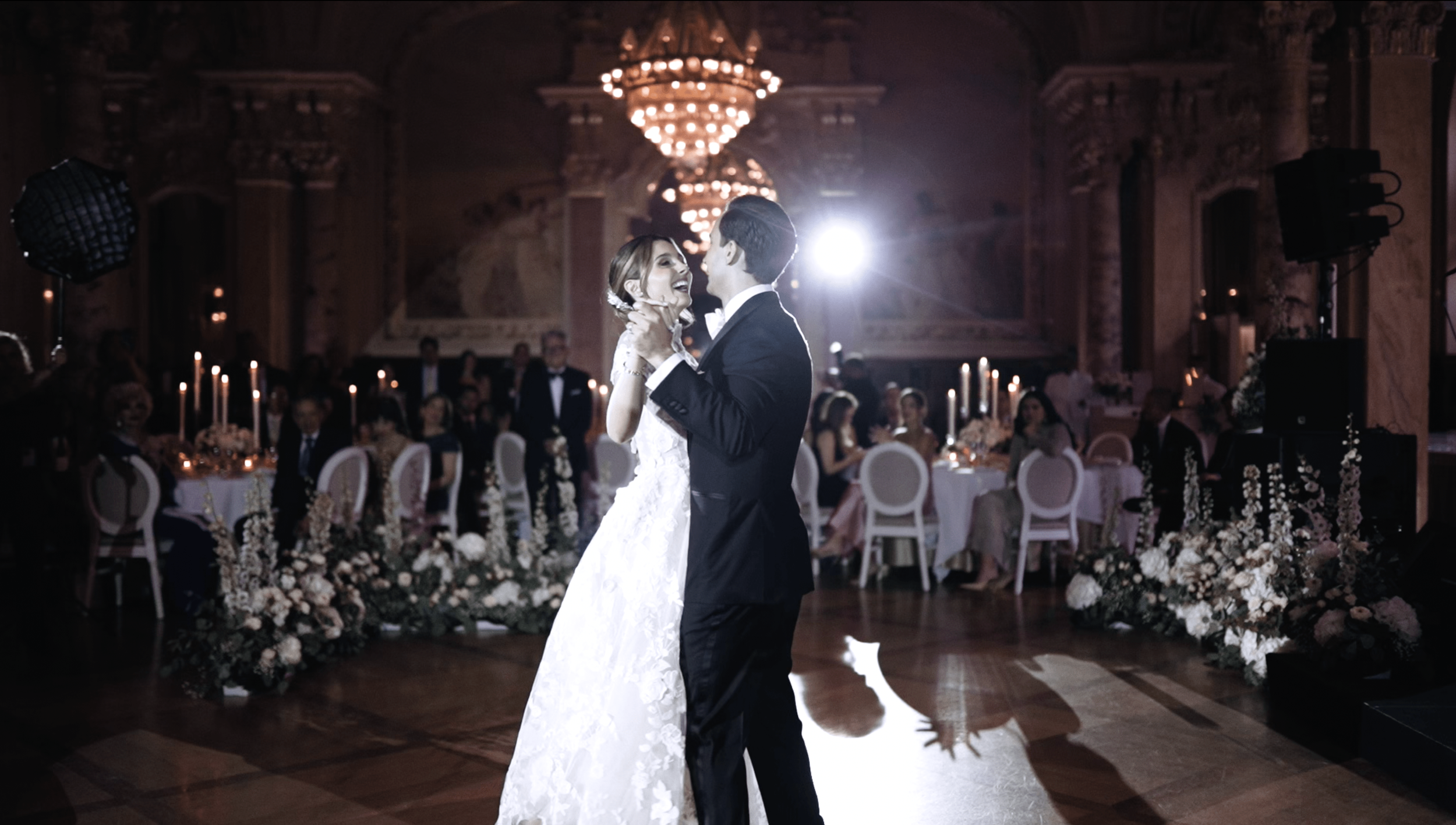 A bride and groom are dancing at their wedding reception in an elegant, decorated ballroom with chandeliers and floral arrangements.