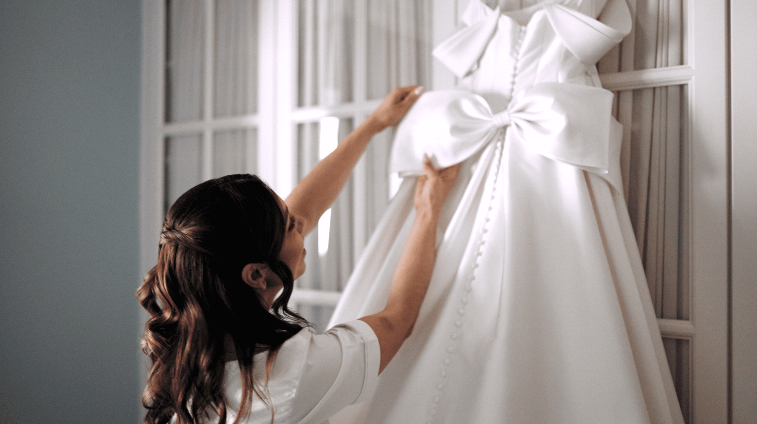 A woman with long, brown hair arranging a white wedding dress with a large bow on the back, hanging on a wall.