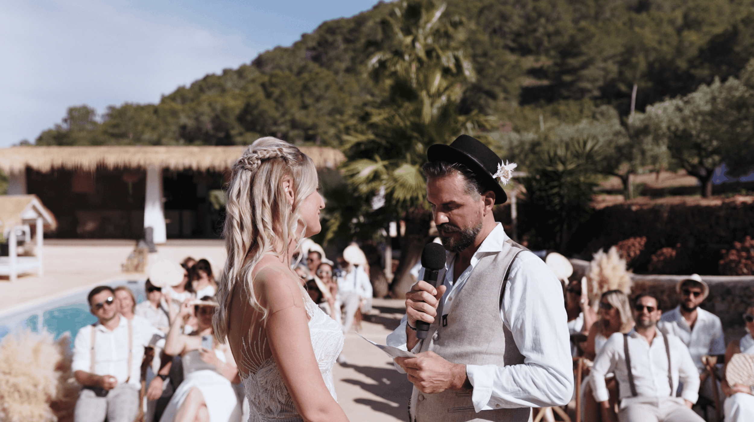 A wedding ceremony outdoors with the bride and groom exchanging vows, surrounded by seated guests on a sunny day with trees and a rustic building in the background.