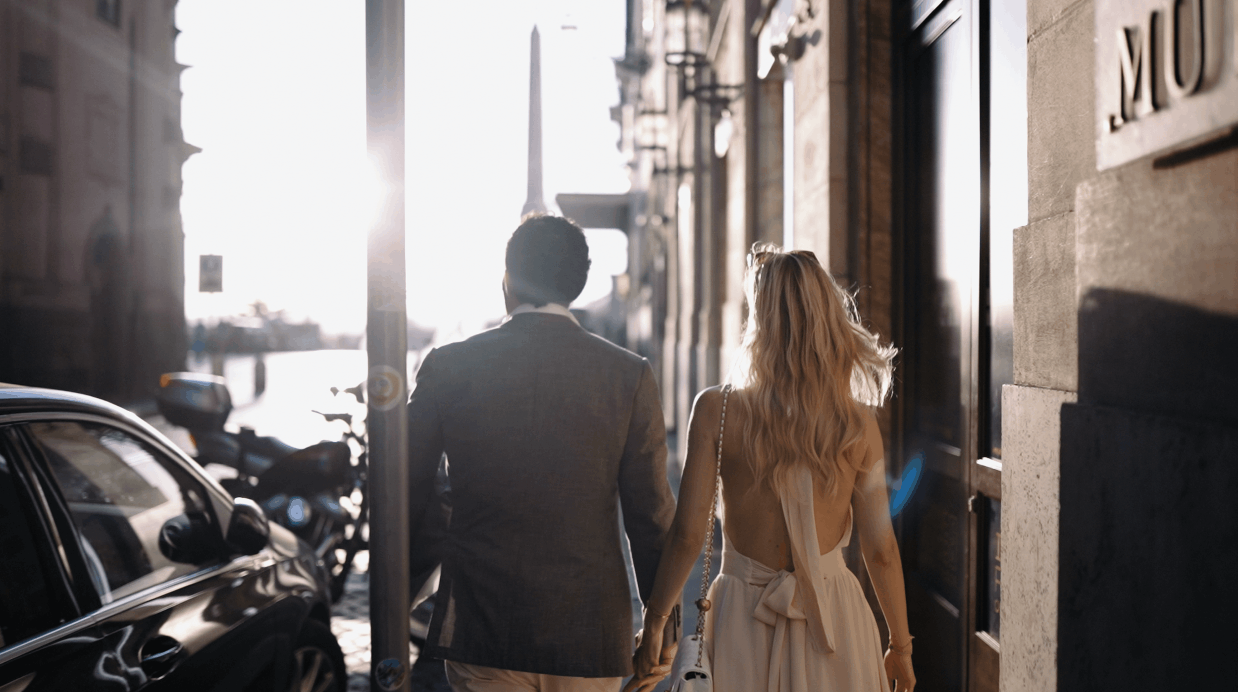 A man and woman walking hand in hand on a city sidewalk during sunset, with parked cars and a building in the background.