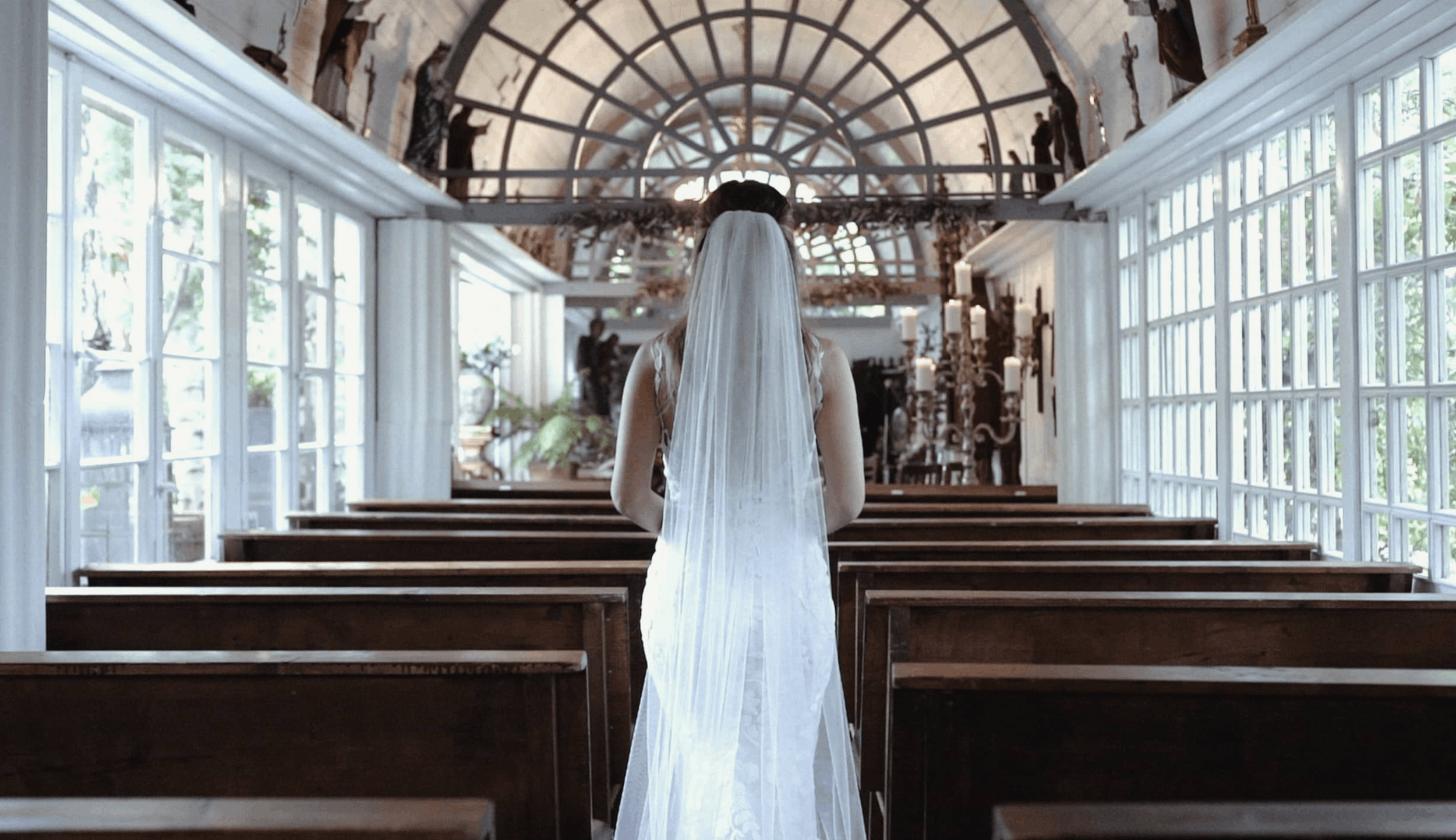 A woman in a wedding dress and veil standing from behind inside a church with wooden pews and large windows, facing an altar with candles and statues.