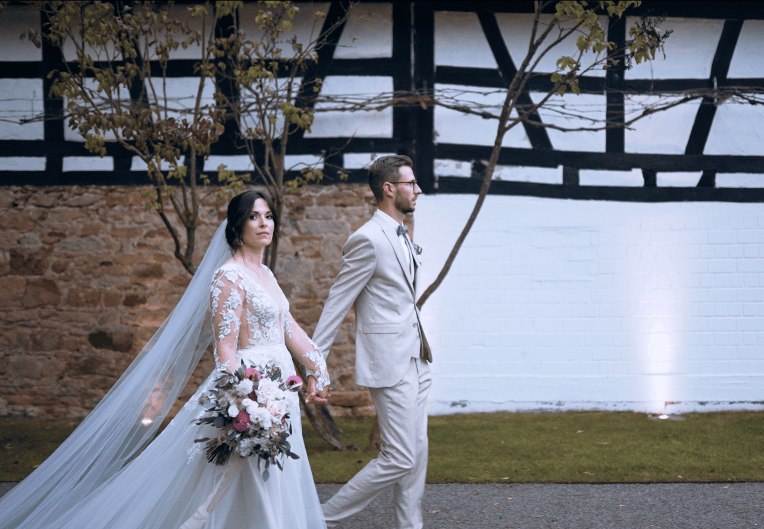 A bride and groom holding hands during a wedding ceremony outdoors. The bride wears a white lace wedding dress with a long veil, and the groom wears a light-colored suit with a bow tie. The bride is holding a bouquet of flowers, and they are standing in front of a rustic brick and wood building with trees nearby.