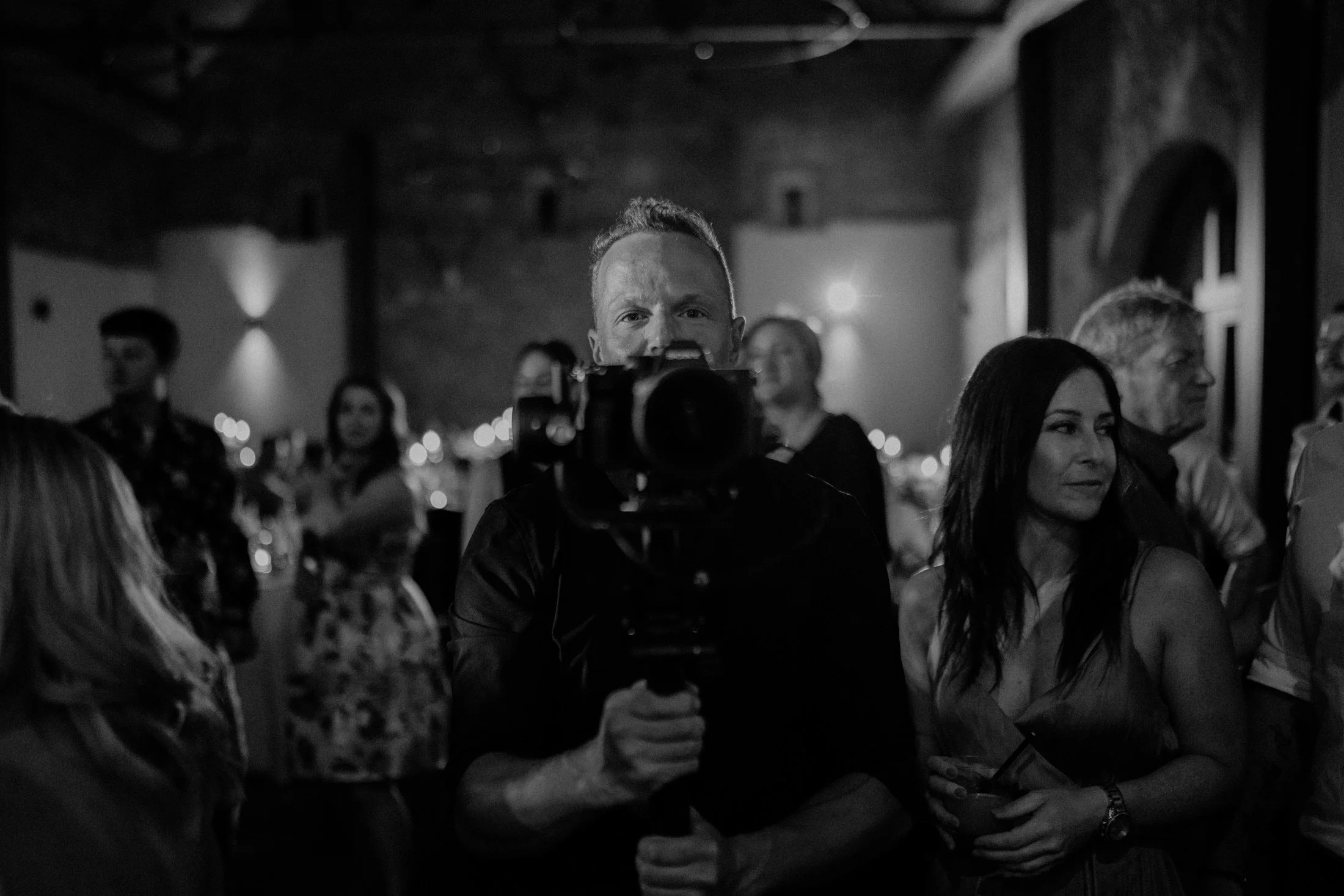 Black-and-white photo of a man with light skin, short hair, and a T-shirt, looking directly into the camera. He is indoors at a social gathering with other people, some of whom are women. The background shows dim lighting and arched windows, suggesting a bar or restaurant setting.