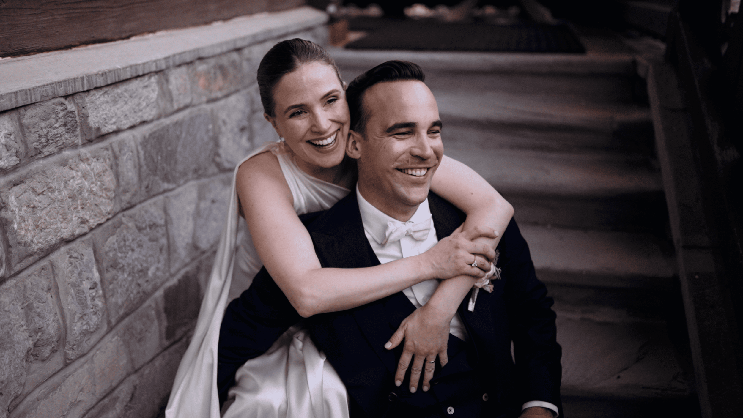 A joyful bride and groom sit on stairs, with the bride embracing the groom from behind, both smiling happily. The bride has her arms around the groom's shoulders and is wearing a white dress, while the groom is in a dark tuxedo and white bow tie. They are sitting against a rustic stone wall.