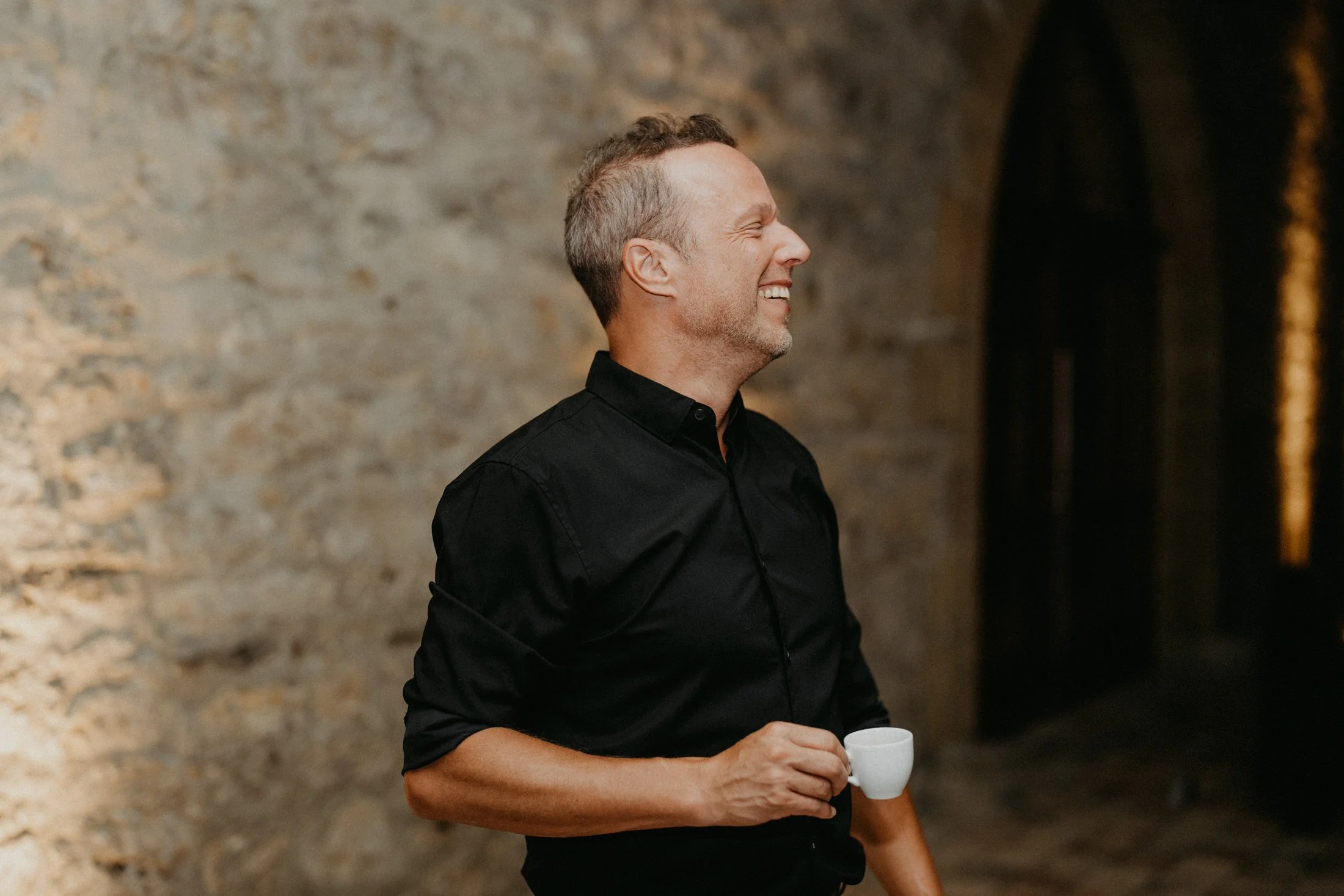 A middle-aged man with short, graying hair, wearing a black shirt, smiling and holding a small white cup, standing against a textured stone wall.