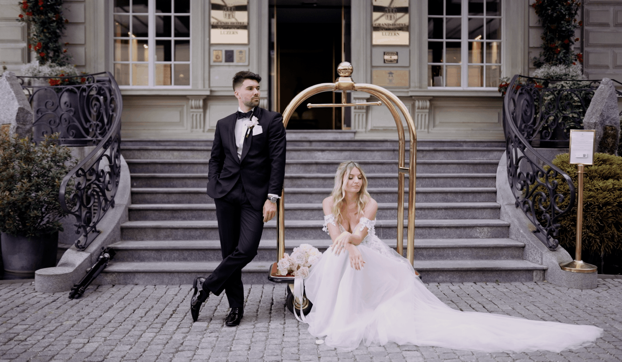 A bride in a white wedding dress sitting on steps in front of a building with large windows, next to a groom in a black tuxedo with a bow tie, standing beside a luggage cart with a bouquet of flowers on it. The scene is decorated with floral arrangements and a brass luggage cart, with a cobblestone street in the foreground.