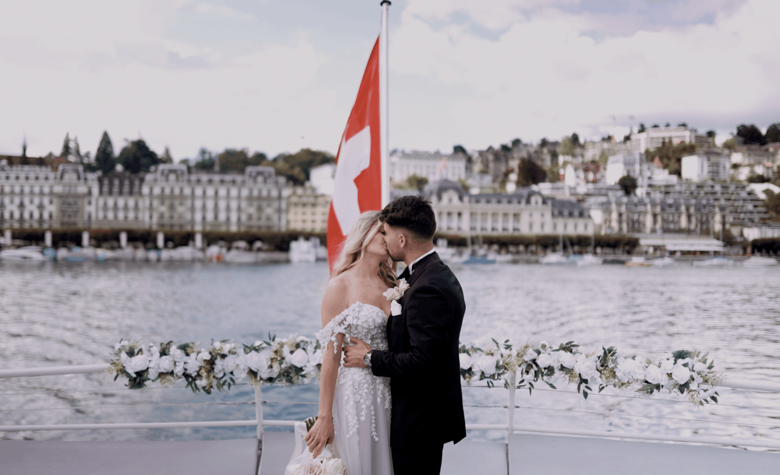A bride and groom kissing on a boat deck by the water, with buildings and hills in the background, and an American flag behind them.